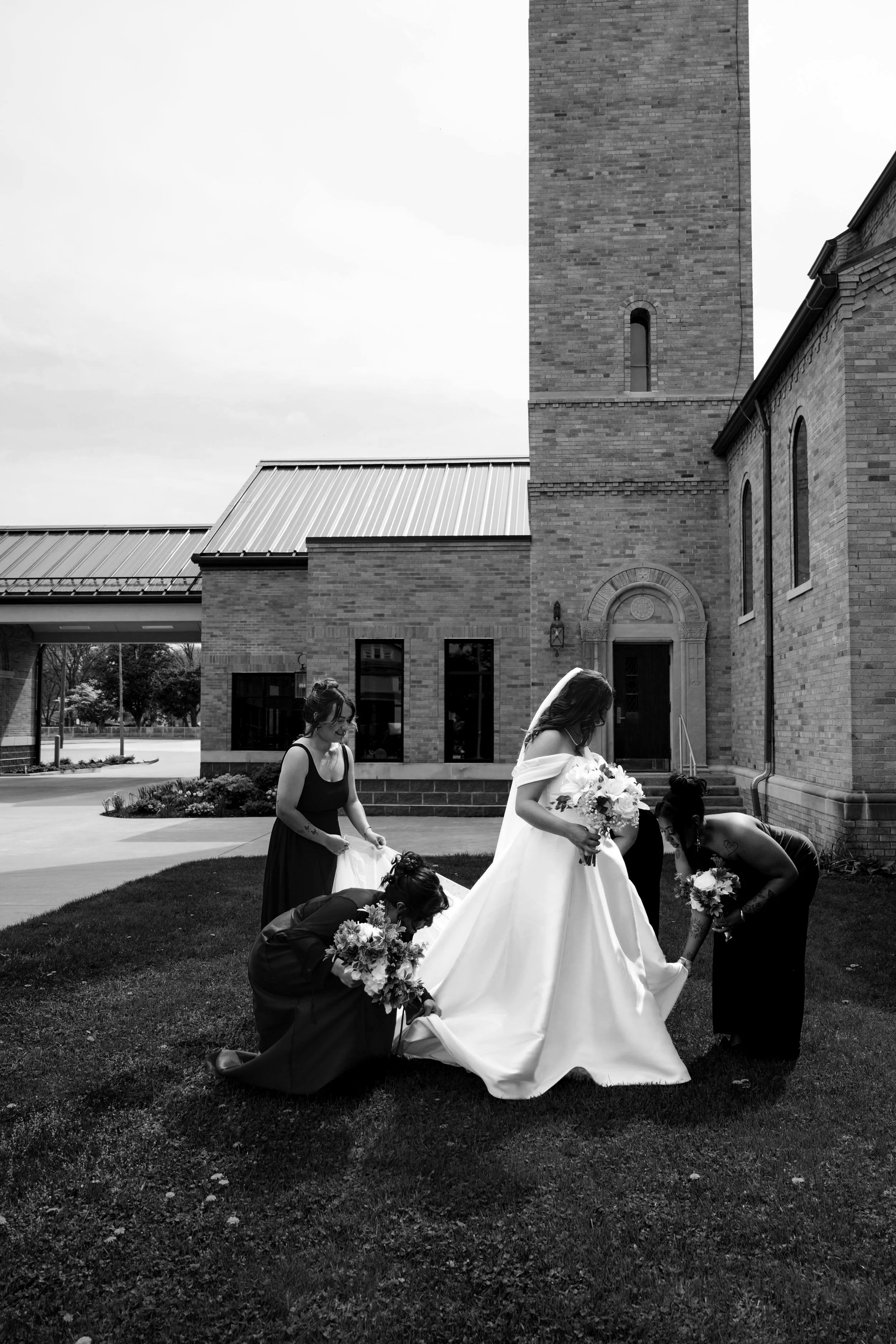 A bride and bridesmaids preparing outside a church for a wedding, with some women adjusting her wedding dress and holding bouquets.