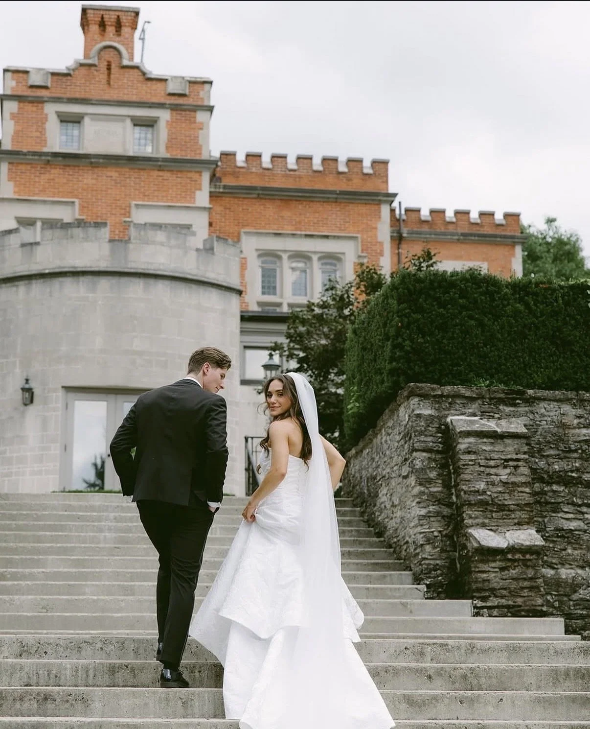 A bride and groom standing on outdoor steps in front of a historic brick building, with the groom looking back at the bride and the bride smiling at the camera.