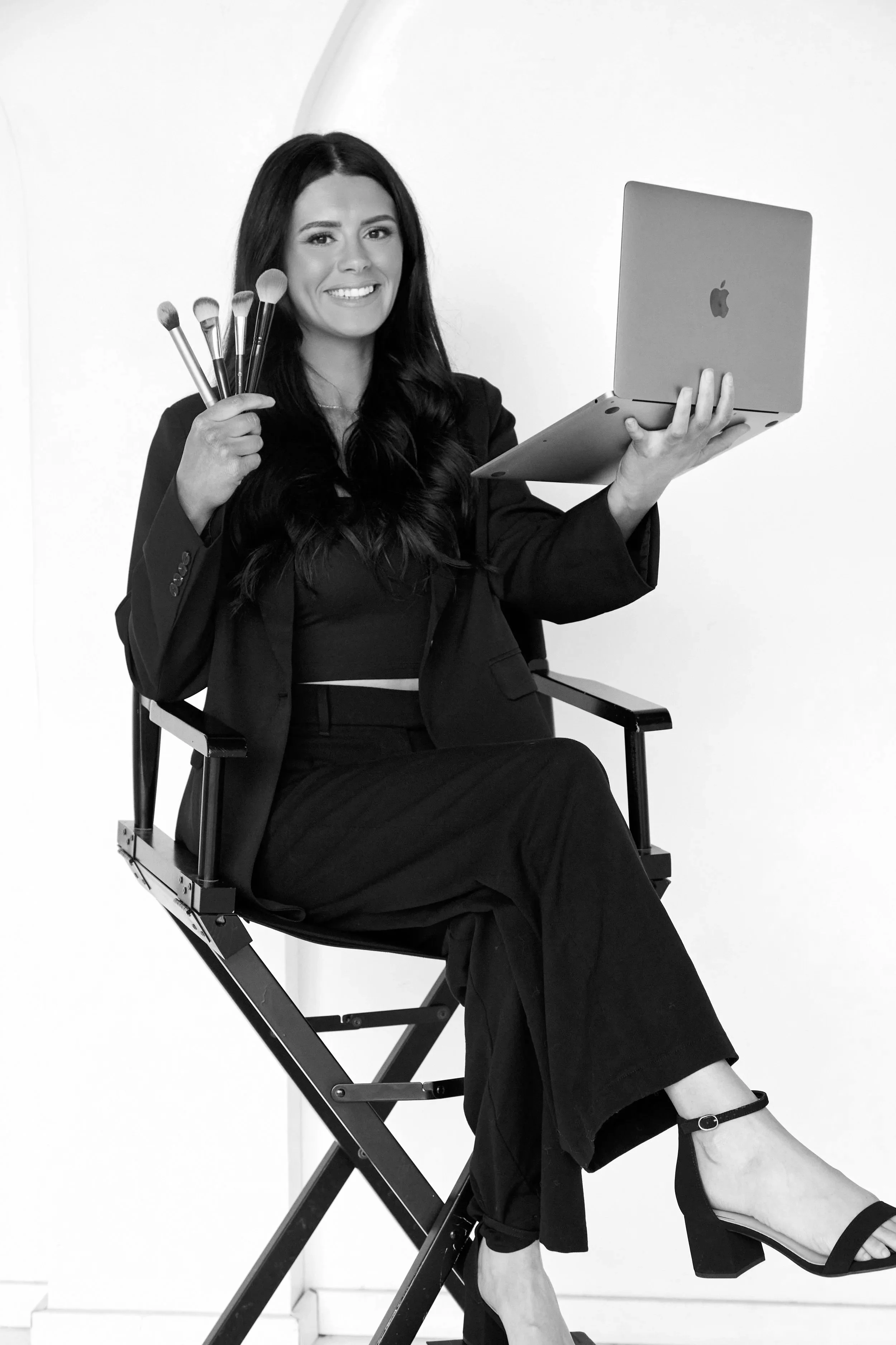 A woman with long dark hair in a black blazer and skirt sitting on a director's chair, holding a laptop in one hand and makeup brushes in the other, smiling at the camera in front of a plain white background.