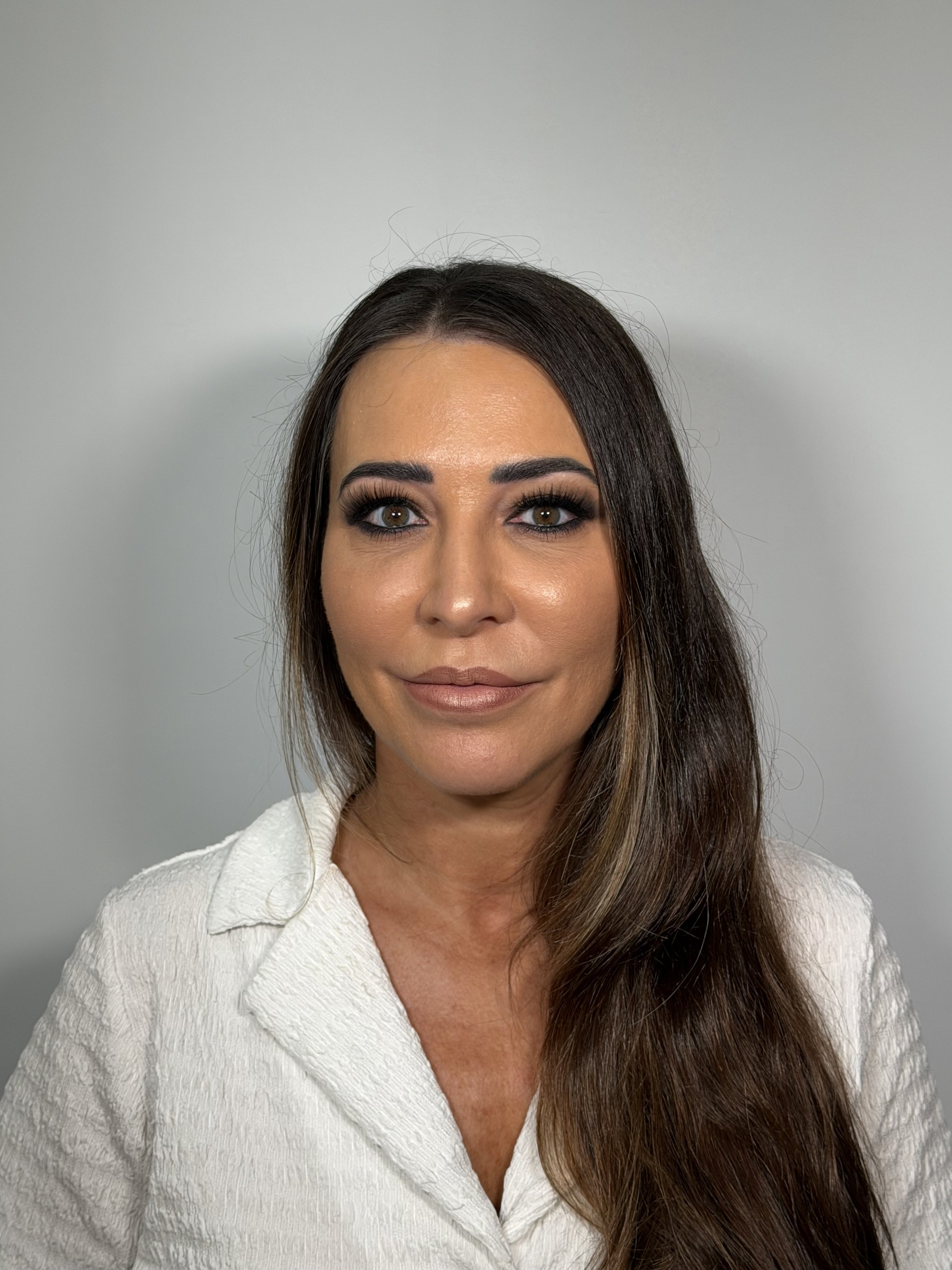 Close-up portrait of a woman with long brown hair, brown eyes, and makeup, wearing a white textured top against a plain gray background.