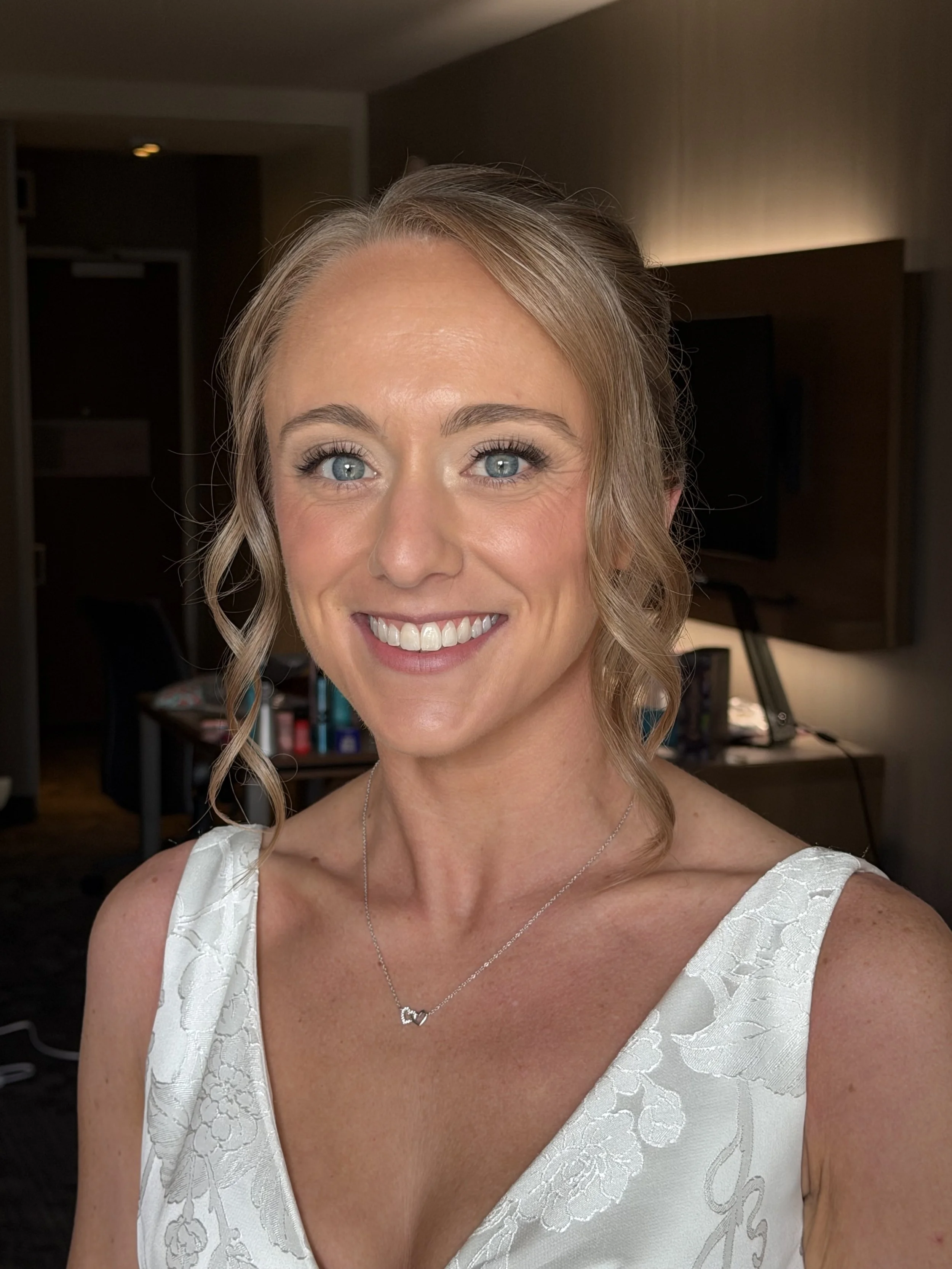 A woman with blonde hair styled in loose curls, wearing a white dress with floral embroidery, and a silver necklace with a heart-shaped pendant, smiling indoors in a room with warm lighting and dark furniture.