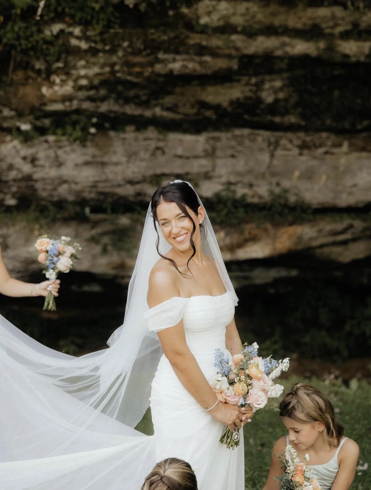 A smiling bride in a white strapless wedding gown holding a bouquet of pastel-colored flowers, standing outdoors with a rock formation in the background, with children and a person holding a bouquet nearby.