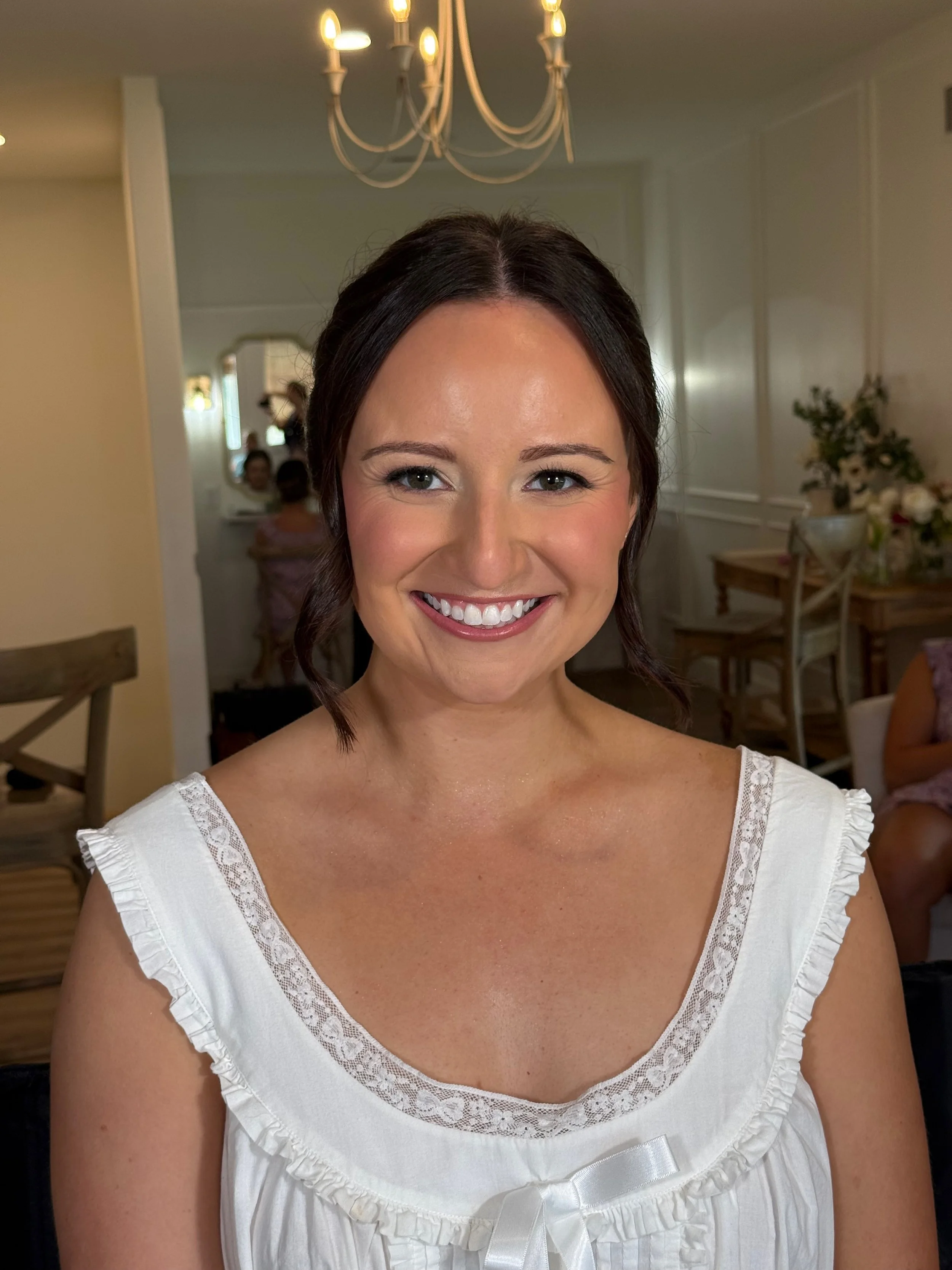 A woman with dark hair styled in loose waves, smiling, wearing a white top with lace trim and a satin bow, standing indoors with a chandelier overhead and blurred background of people and furniture.