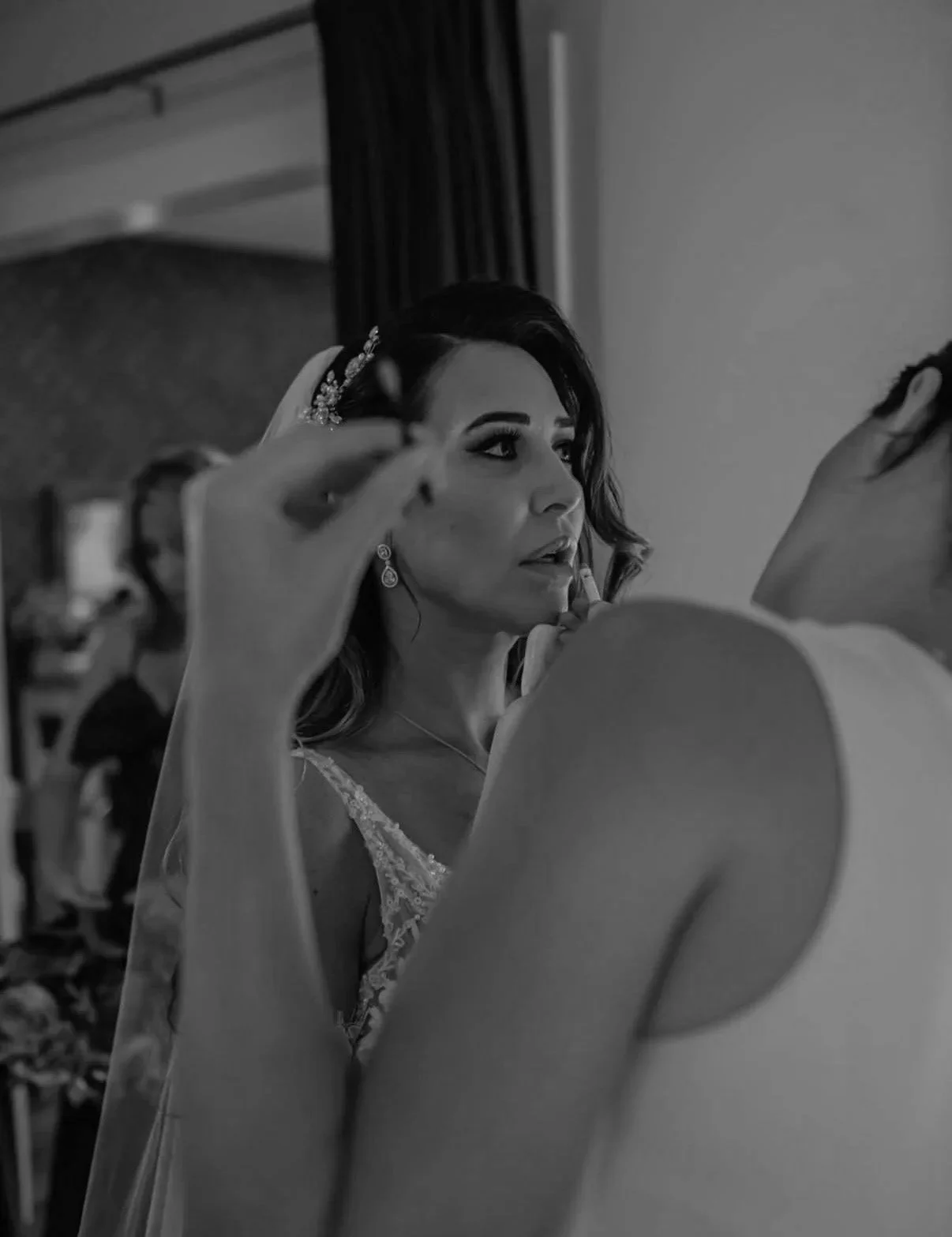 A bride getting her makeup done, seen through a mirror, with a makeup artist applying lipstick, in a room with curtains and a person in the background.