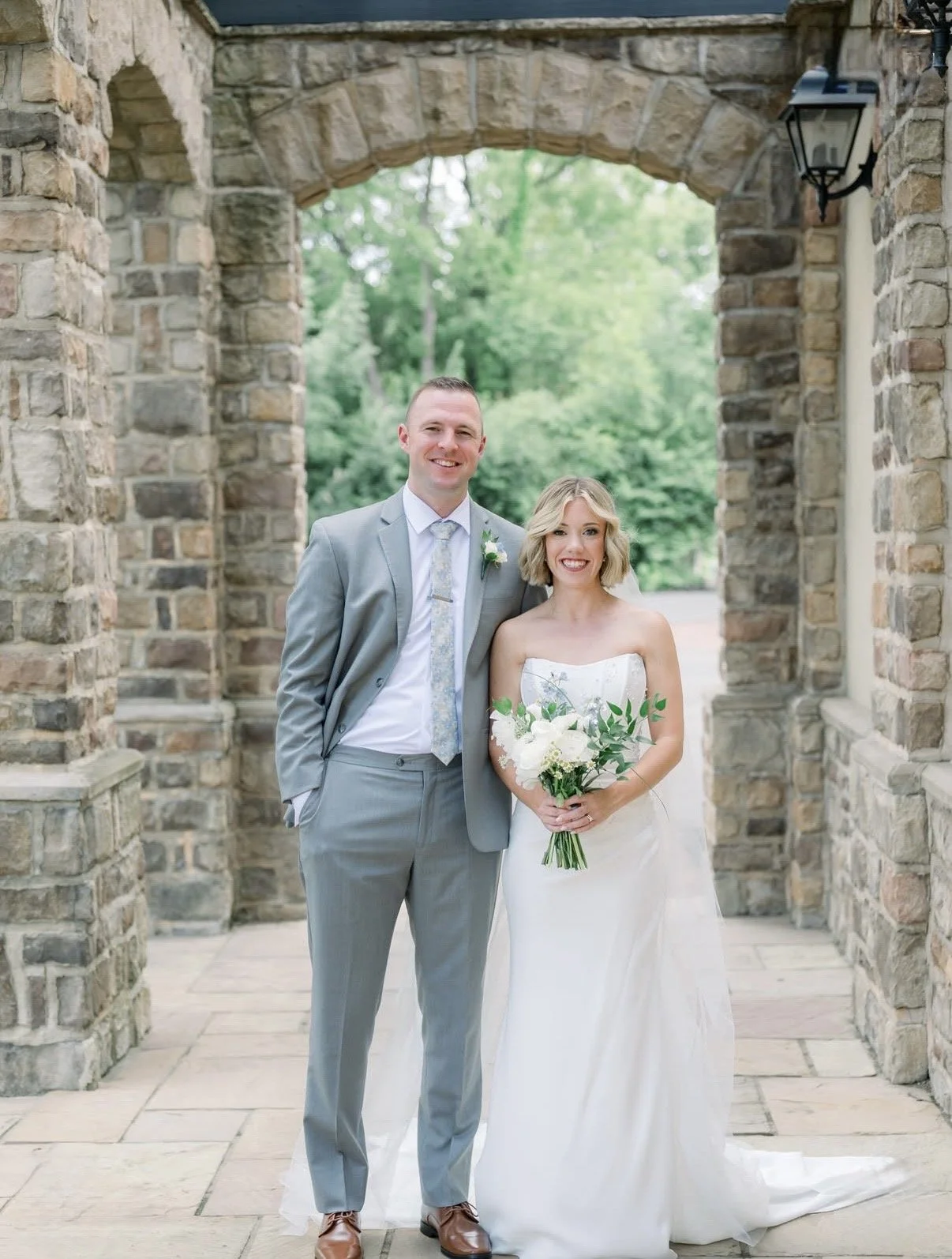 A bride and groom standing together outside under a stone archway, smiling, with the bride holding a bouquet of white flowers and greenery.