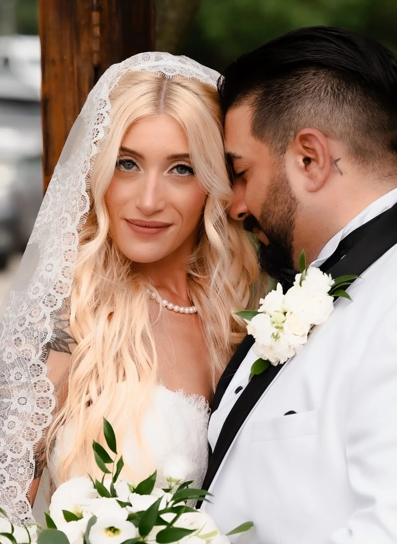 A bride with long blonde hair wearing a lace veil and pearl necklace, standing close to a groom in a white tuxedo with black lapel and boutonniere, both with their heads touching in a tender moment.