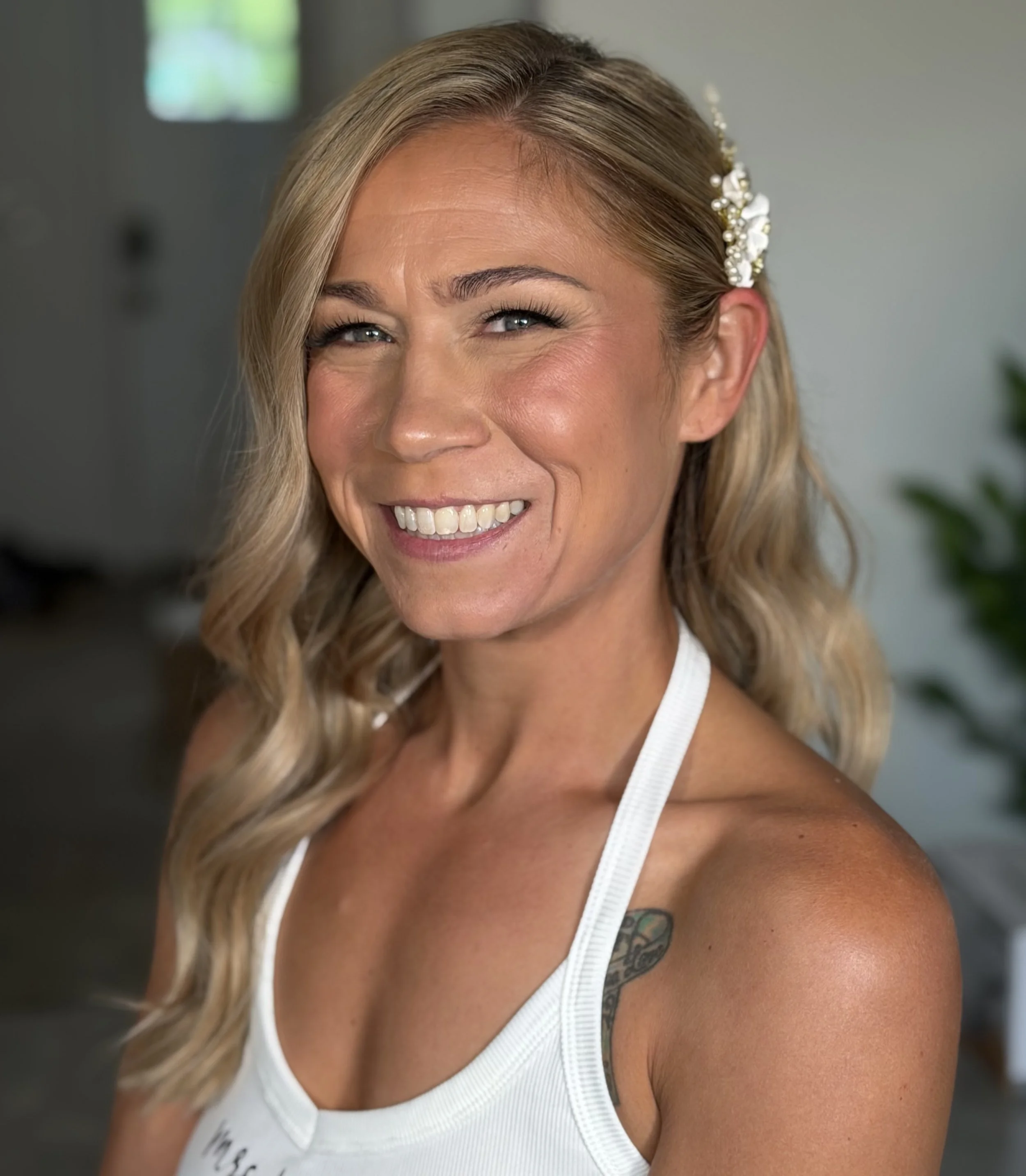 Close-up image of a smiling blonde woman with pearl and flower hair accessory, wearing a white tank top, indoors with blurred background.