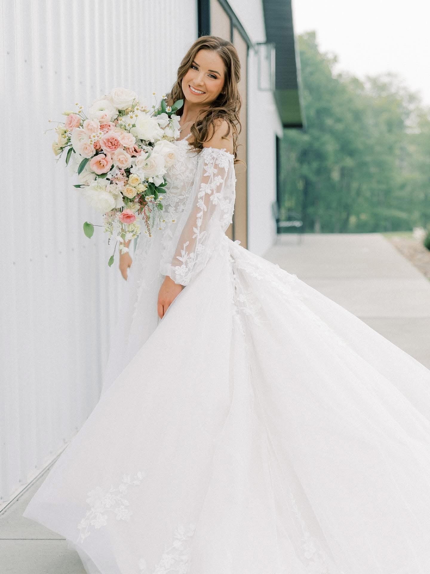 Woman in a wedding dress holding a large bouquet of flowers outside in front of a building with trees in the background.
