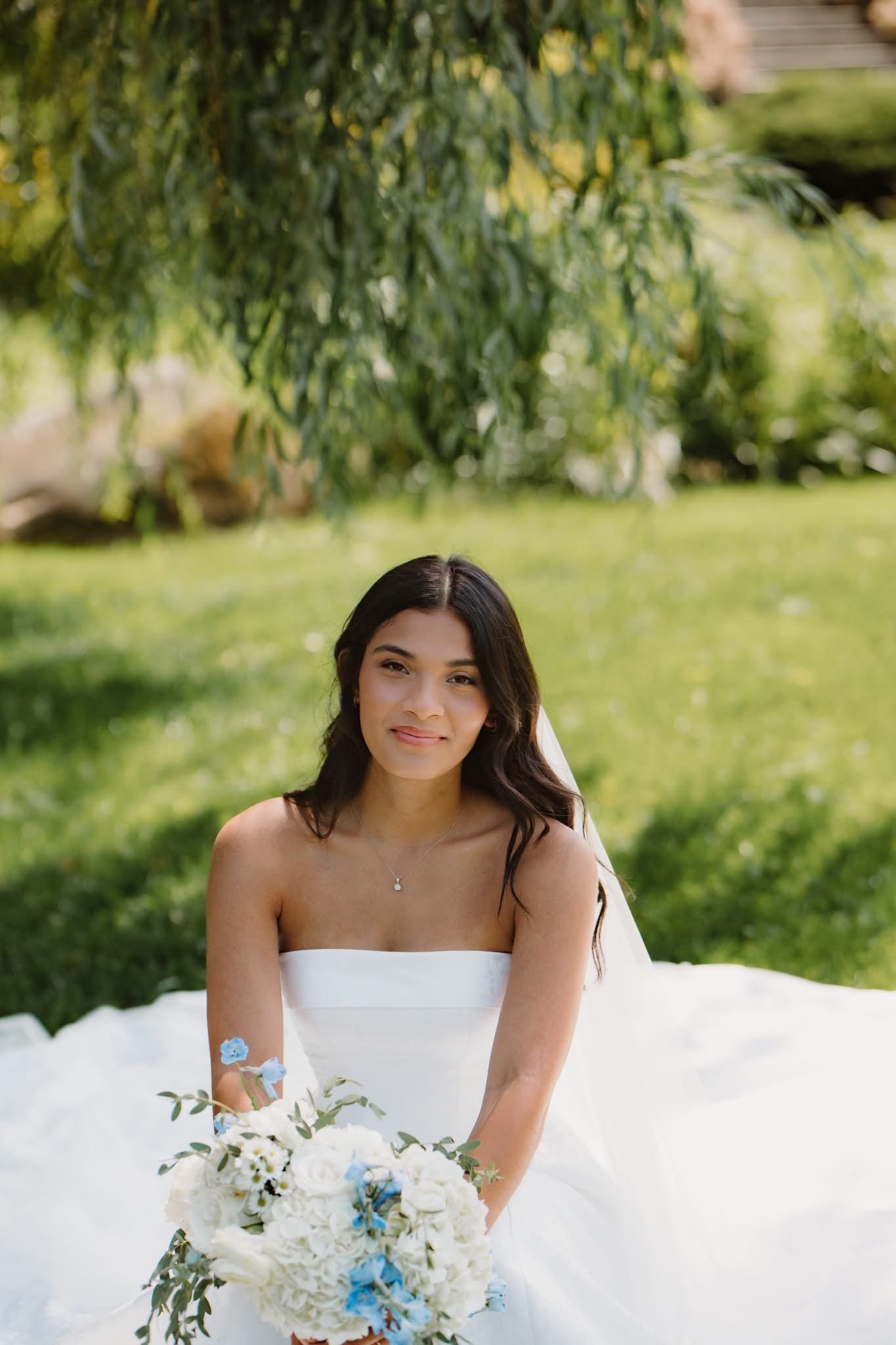 A woman with dark hair sitting outdoors in a white strapless dress, holding a large bouquet of white and blue flowers, with a background of green grass and trees.