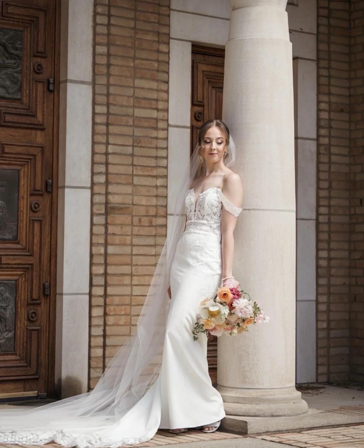 A bride in a white wedding dress with lace details, holding a bouquet of pink, peach, and white flowers, standing outdoors near a brick building with a large white column.