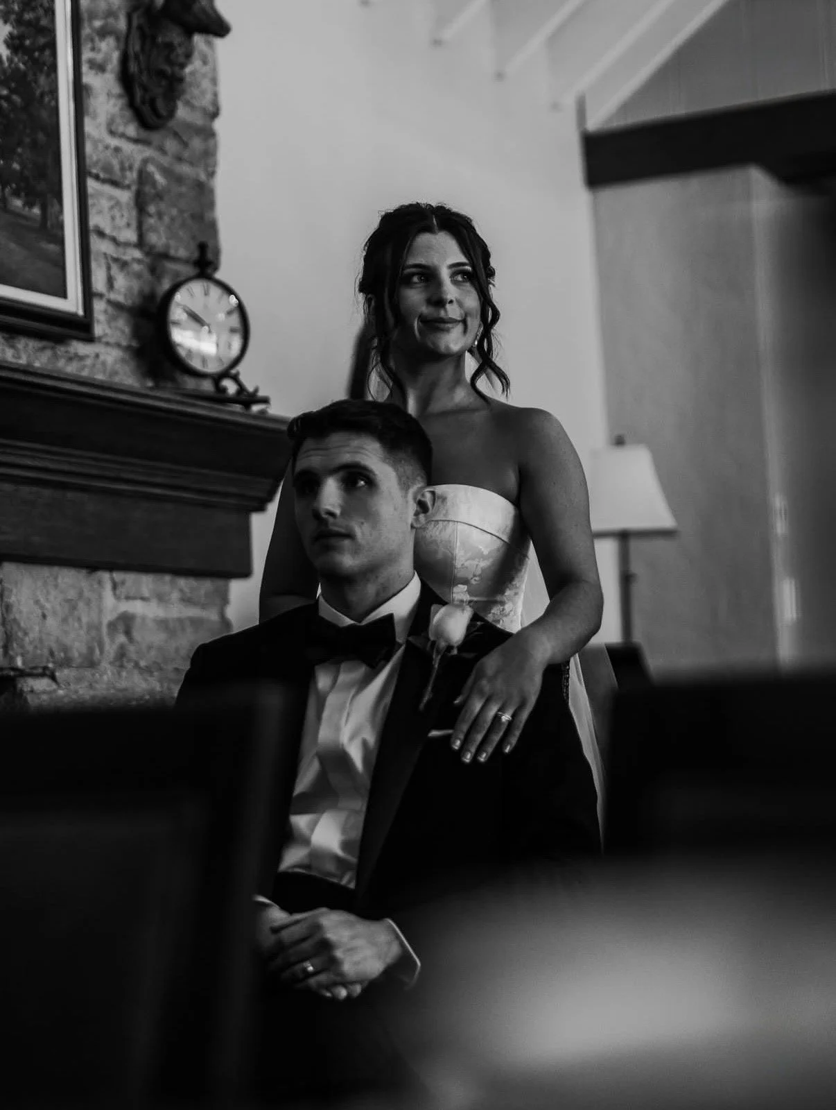 A black-and-white photo of a newlywed couple, the groom kissing the bride on the forehead. The groom is wearing a tuxedo and the bride is in a wedding dress holding a large floral bouquet, with a veil flowing to her right. They are outdoors, with blurred trees in the background.