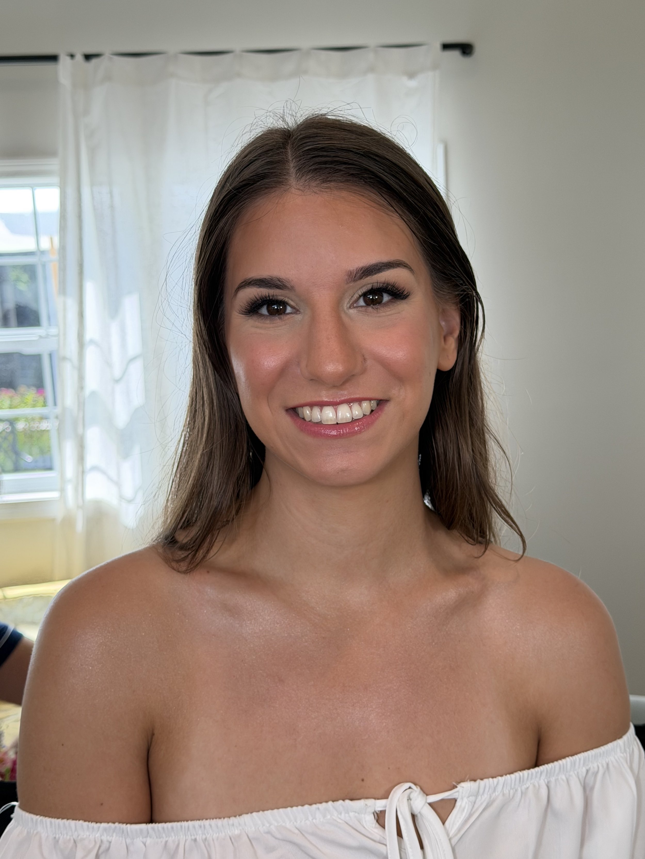 Close-up of a smiling woman with brown hair, wearing an off-the-shoulder white top, inside a room with white curtains and a window in the background.