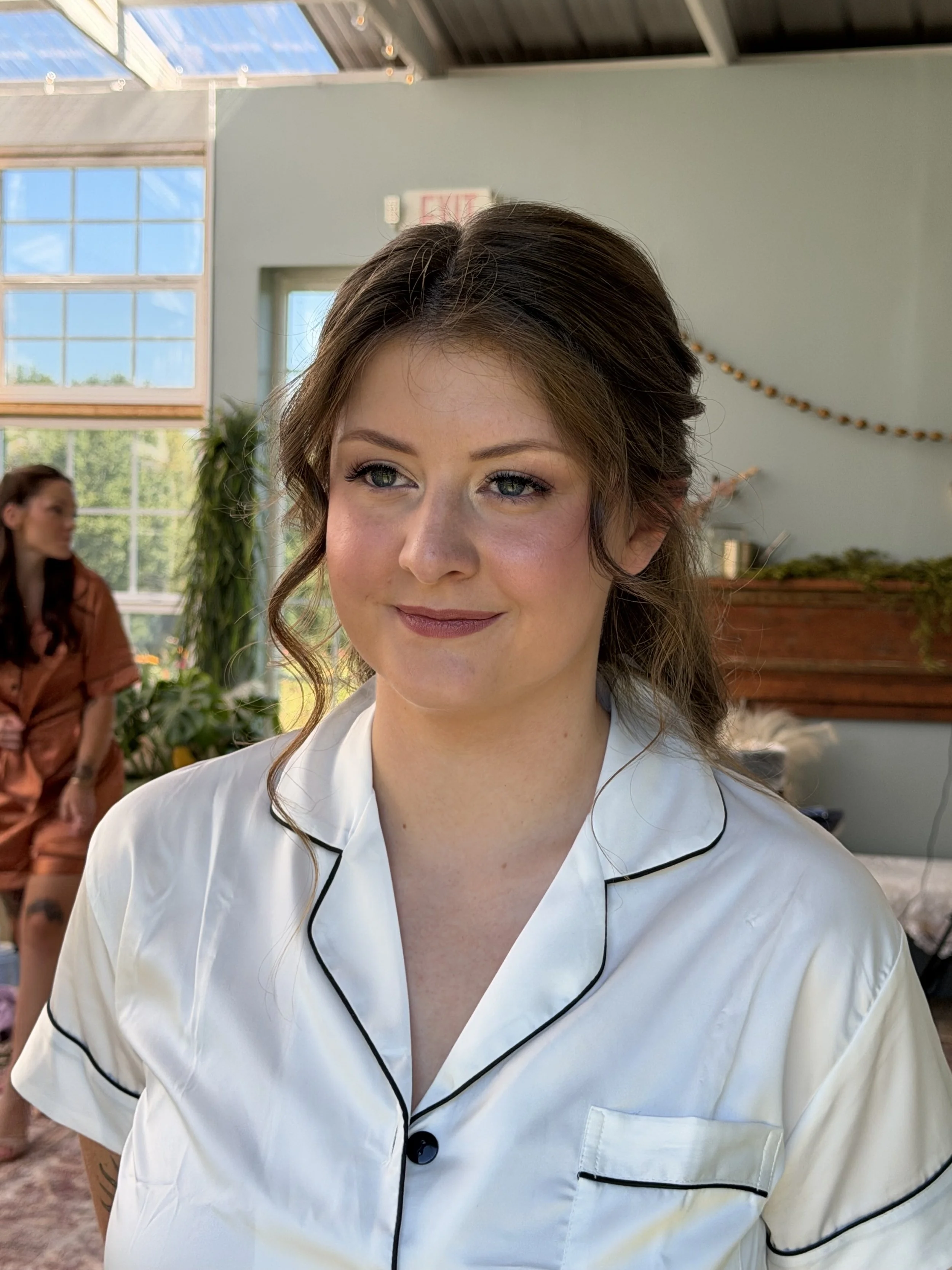 Close-up of a woman with styled brown hair and light makeup, wearing a white satin pajama top with black piping, smiling softly indoors with a woman in an orange outfit and plants in the background.