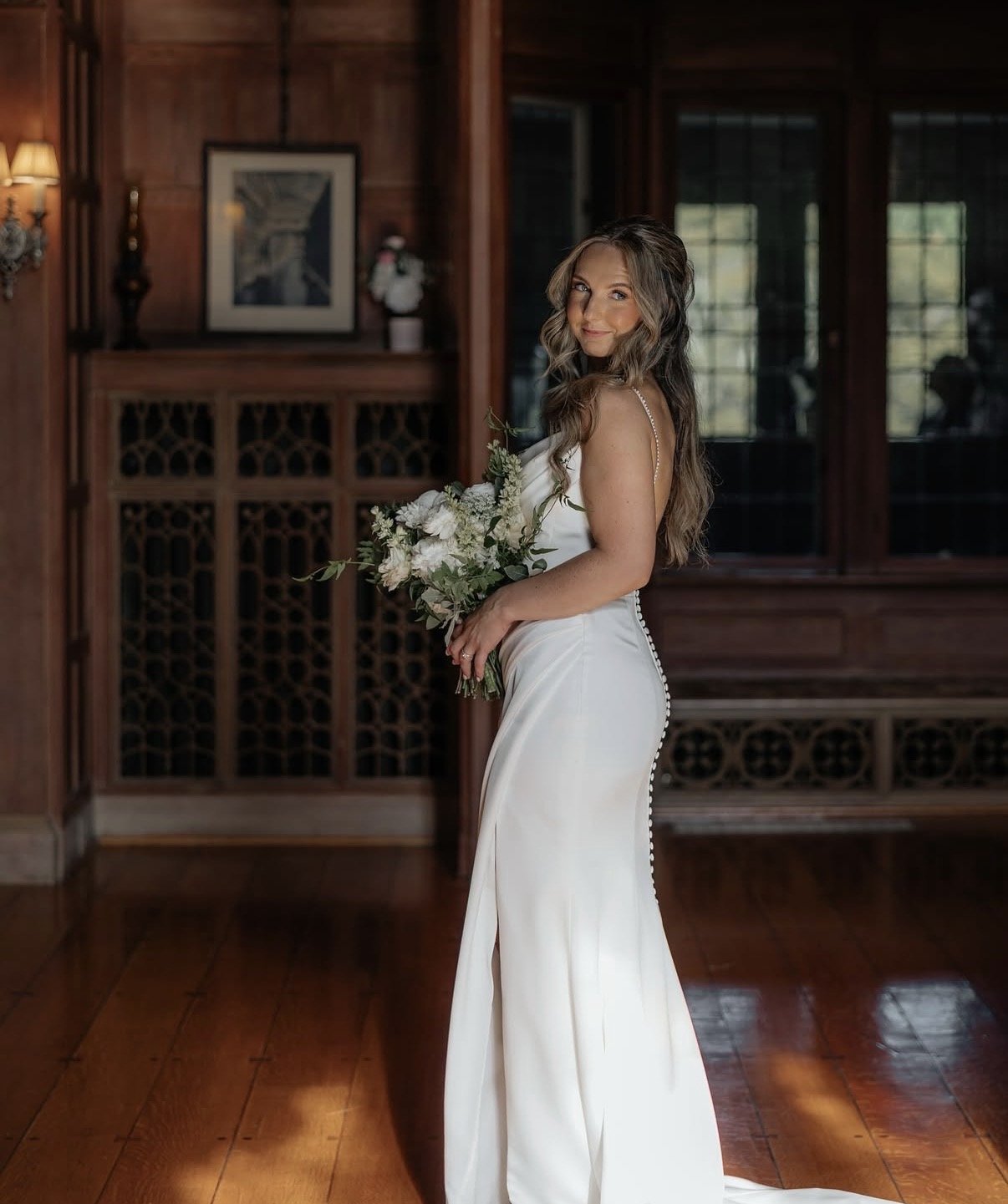 A woman in a white wedding dress holding a bouquet of flowers, standing in a wooden-paneled room with windows and framed artwork.