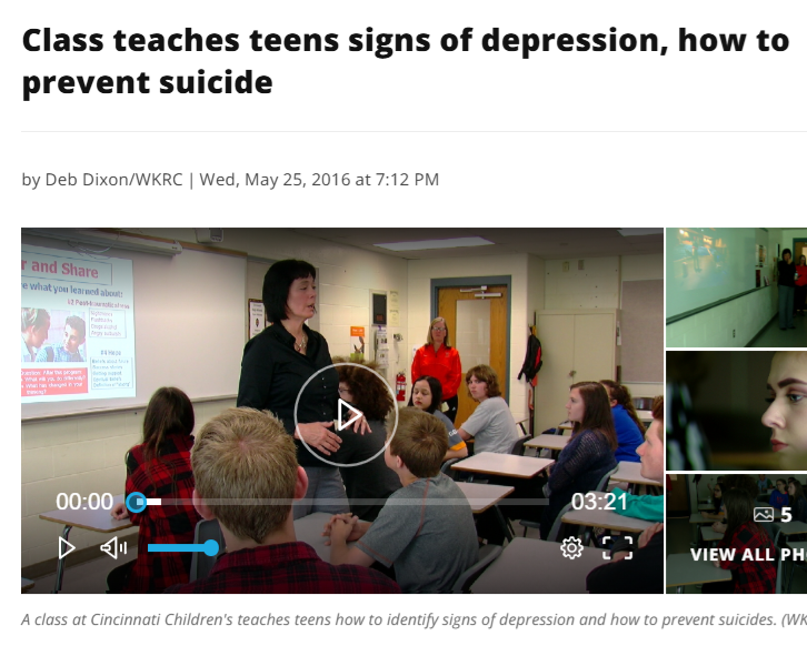 Teacher standing at the front of a classroom teaching teens about recognizing signs of depression and preventing suicides, with students sitting at desks and a projector screen showing information.