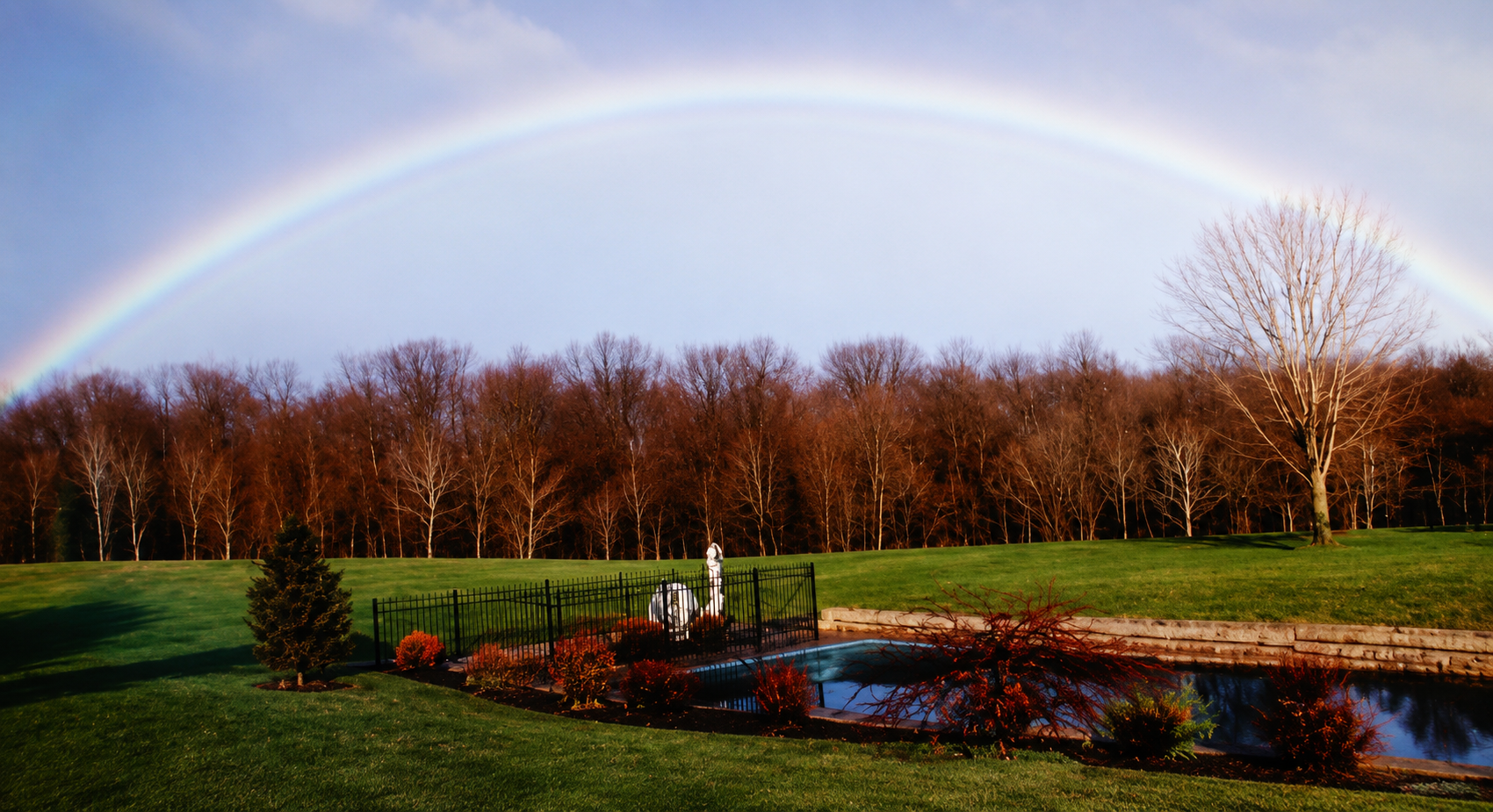 A vibrant rainbow arching across a partly cloudy sky over a landscape with bare trees, green grass, a small pond, and a black metal fence.