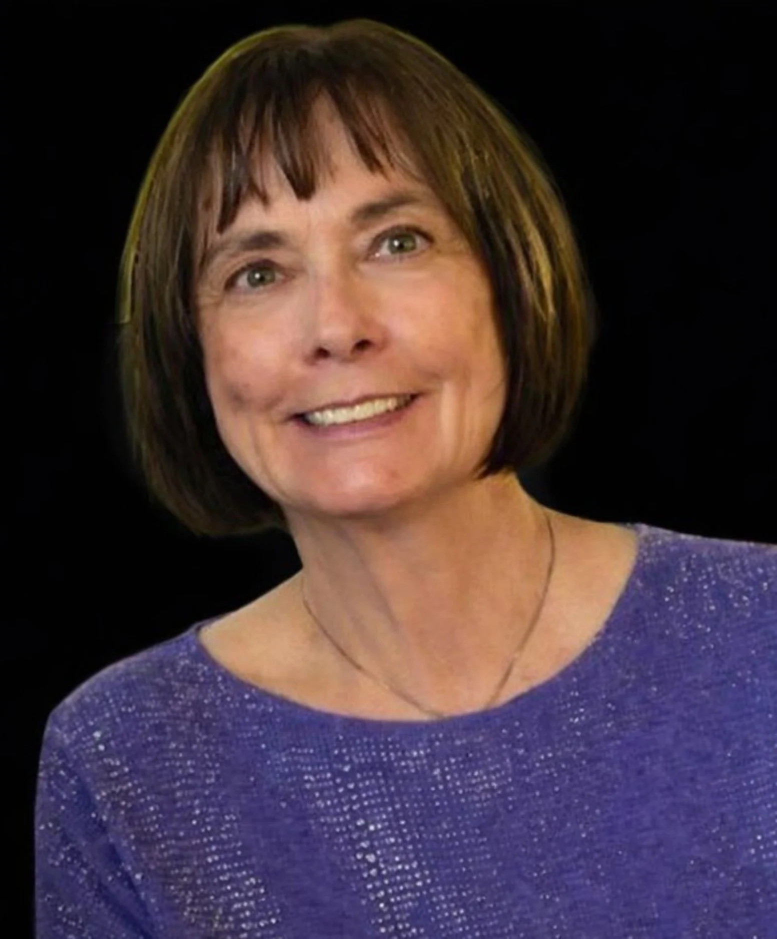 Close-up portrait of a woman with short brown hair, wearing a purple sparkly dress, smiling against a black background.