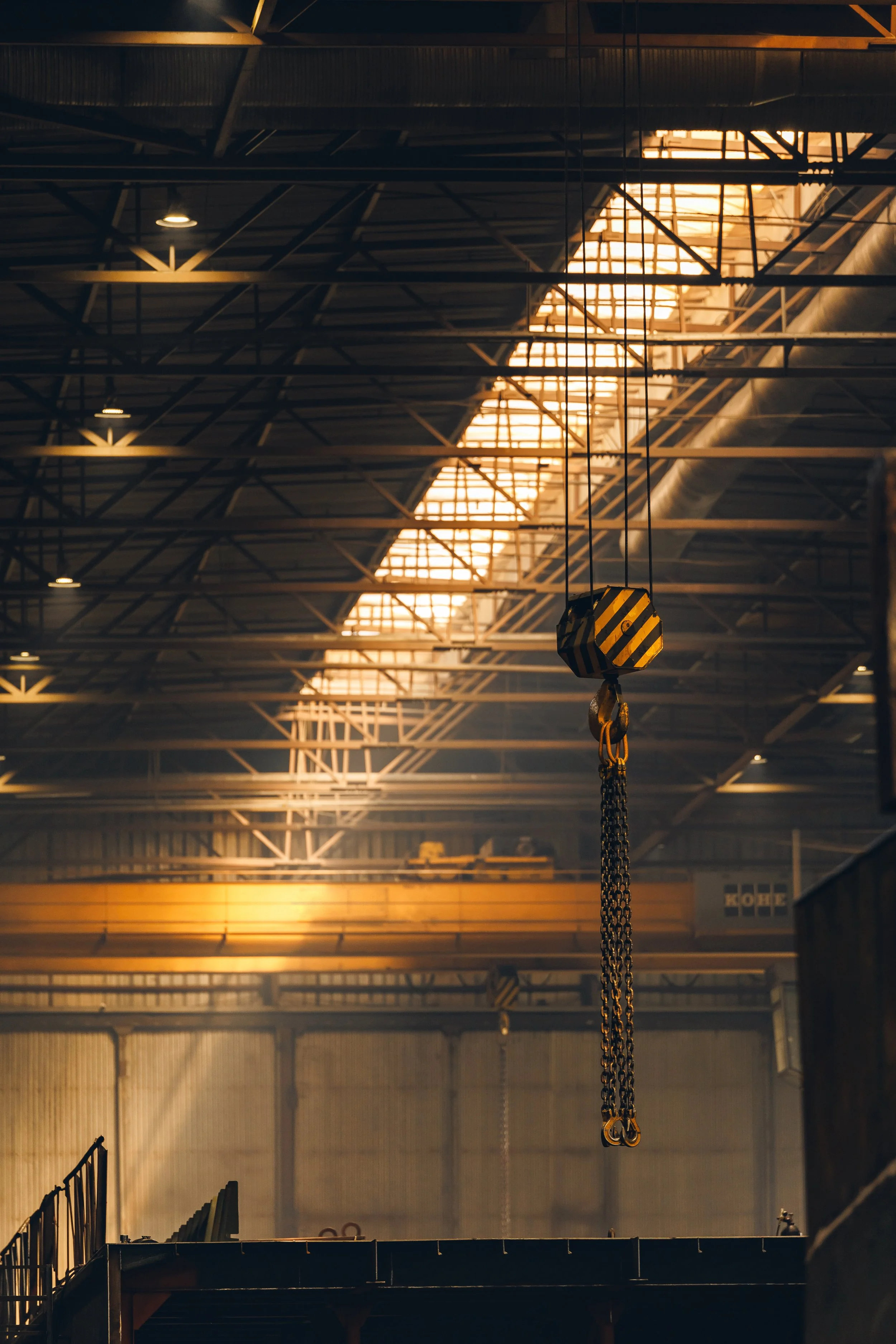 Interior view of an industrial warehouse with a high ceiling, metal beams, and a suspended crane hook hanging from chains.