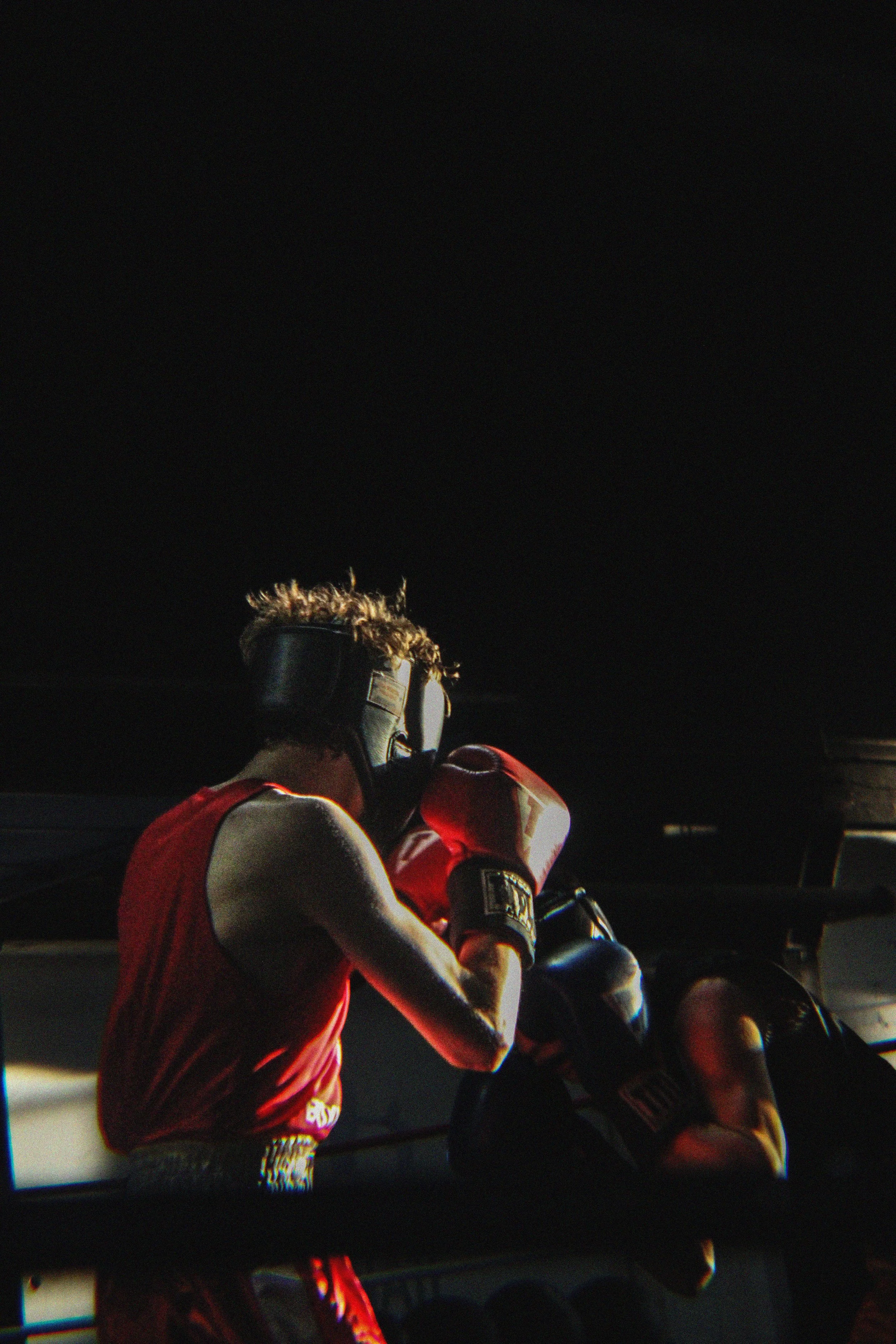 A boxer wearing red gloves and a red jersey with black accents is training. He is wearing a black headgear and has light brown, curly hair. The background is dark with minimal lighting, creating a dramatic effect.