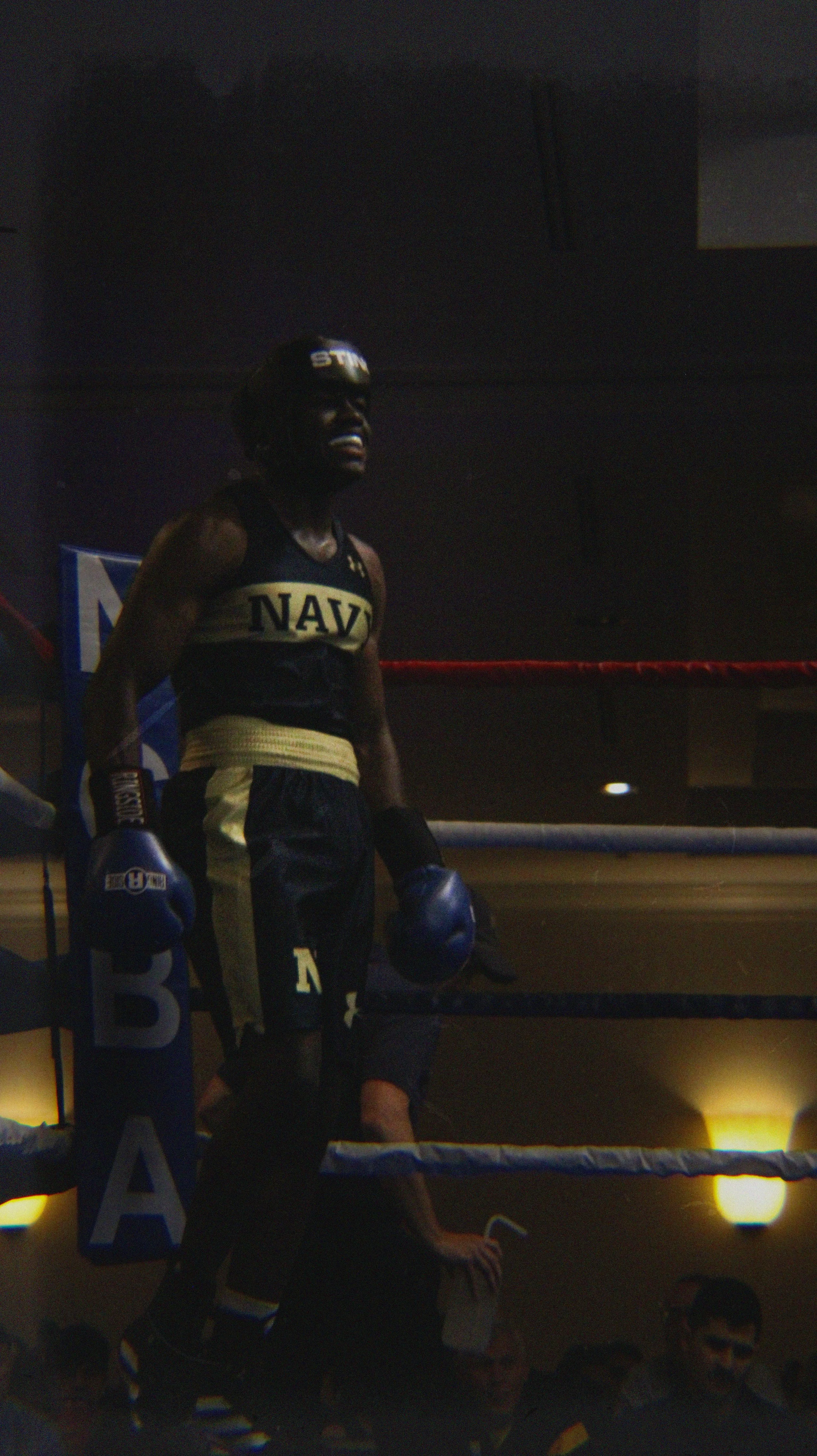 A female boxer in a boxing ring wearing a black and gold uniform with "NAVY" on the chest, blue gloves, and protective headgear. She appears to be tired or exhausted, standing in a ready position.