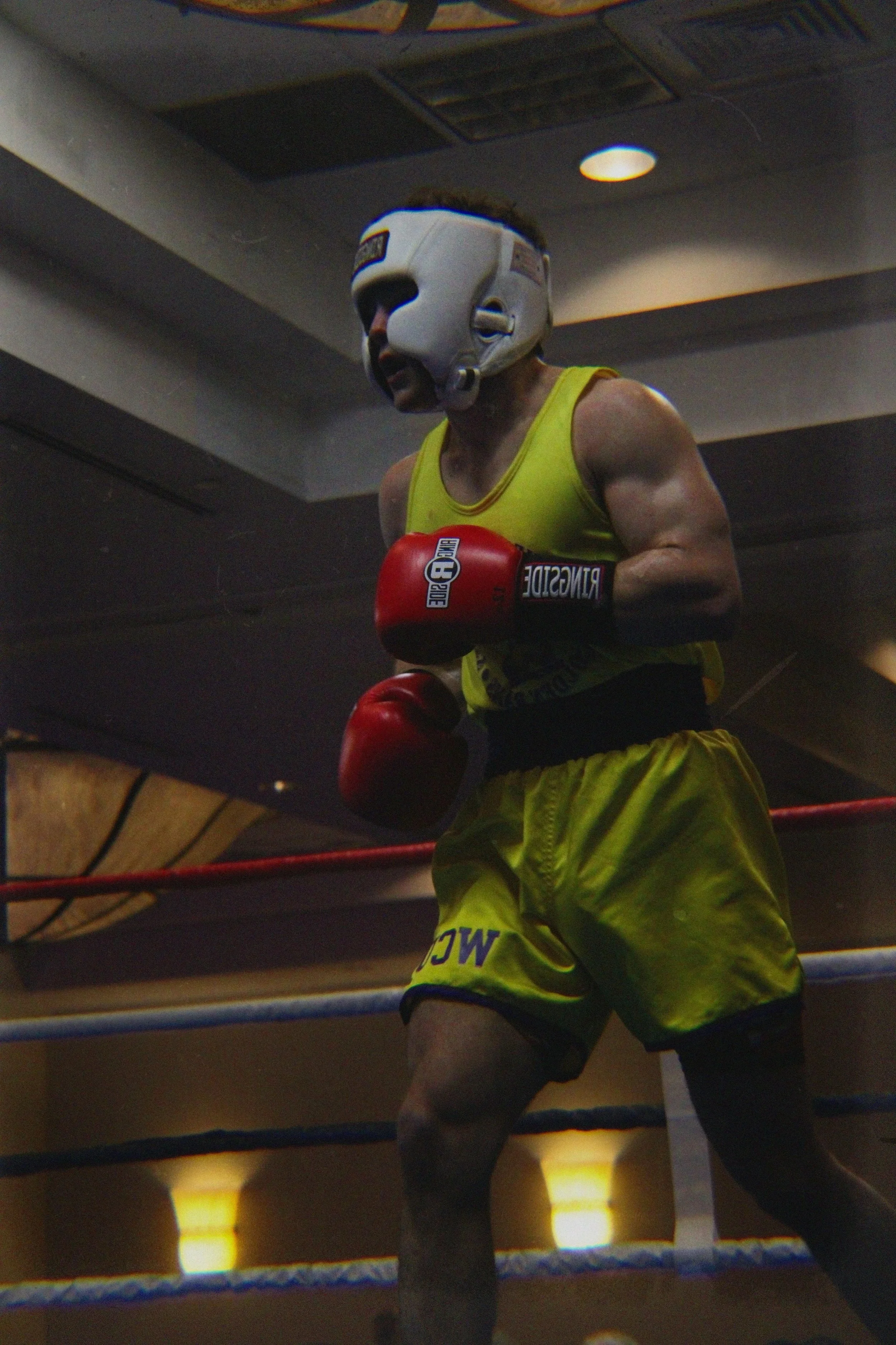 A person wearing a mask resembling a robotic or animal face, yellow boxing shorts, and red gloves is inside a boxing ring. The background shows the ceiling and some lighting of the gym.