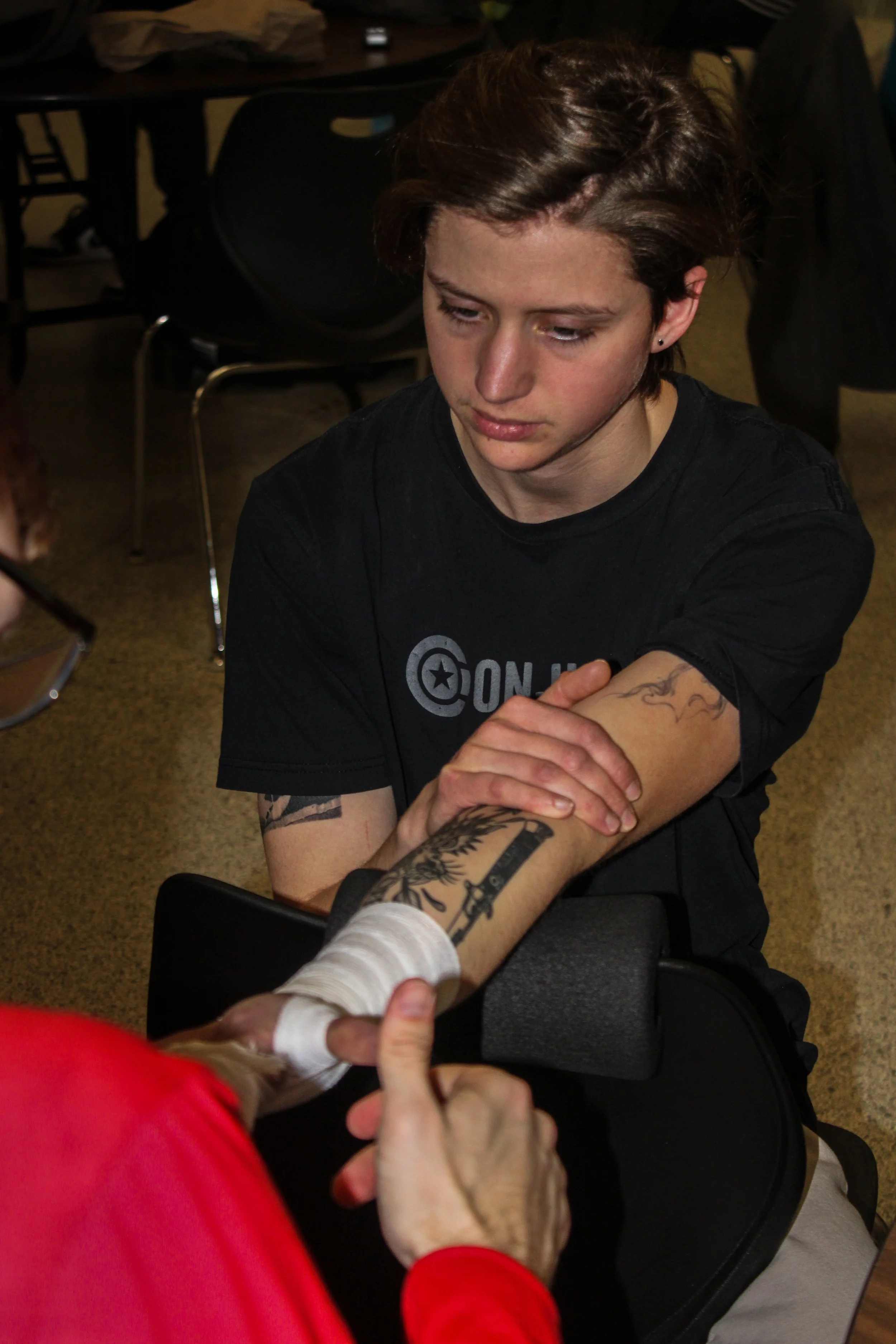 A young person with short brown hair and earrings is sitting and touching the hand of another person, whose hand and tattoos are visible. The setting appears to be indoors, possibly in a public or community space.
