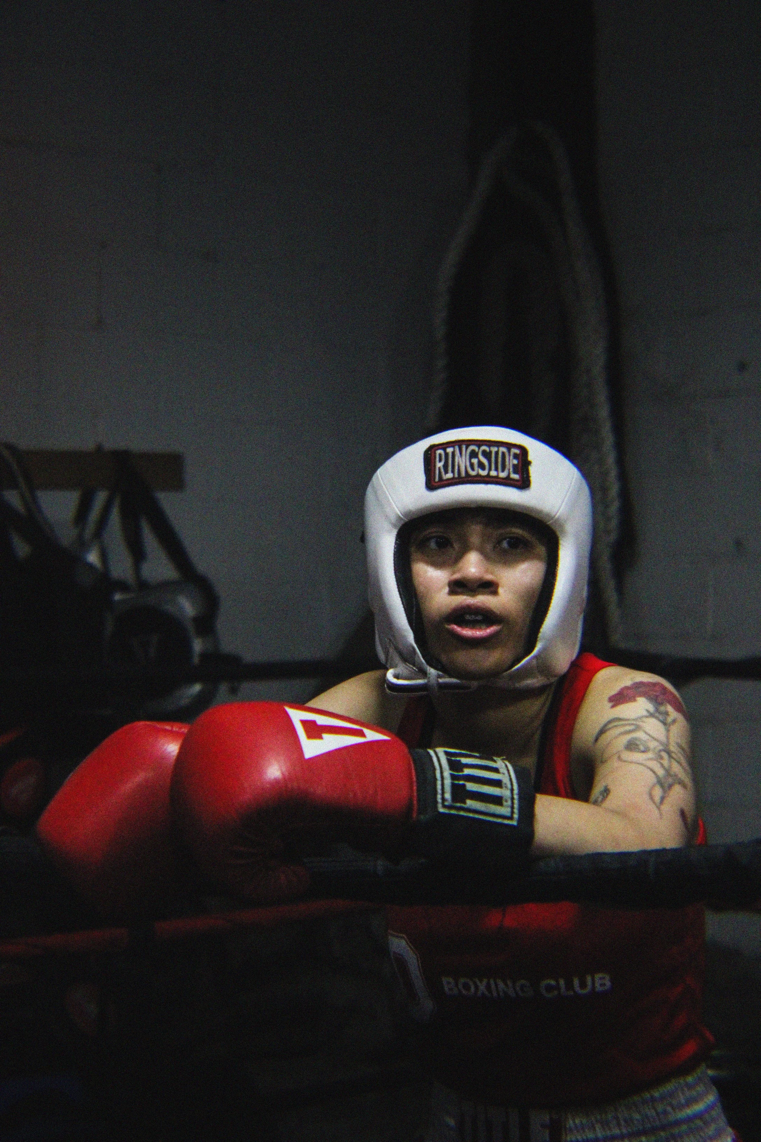 A young boxer wearing a white headgear with the word 'RINGSIDE' on the front, red boxing gloves, and a red and black boxing outfit, sitting with arms crossed inside a boxing gym. The background is dark with gym equipment visible.