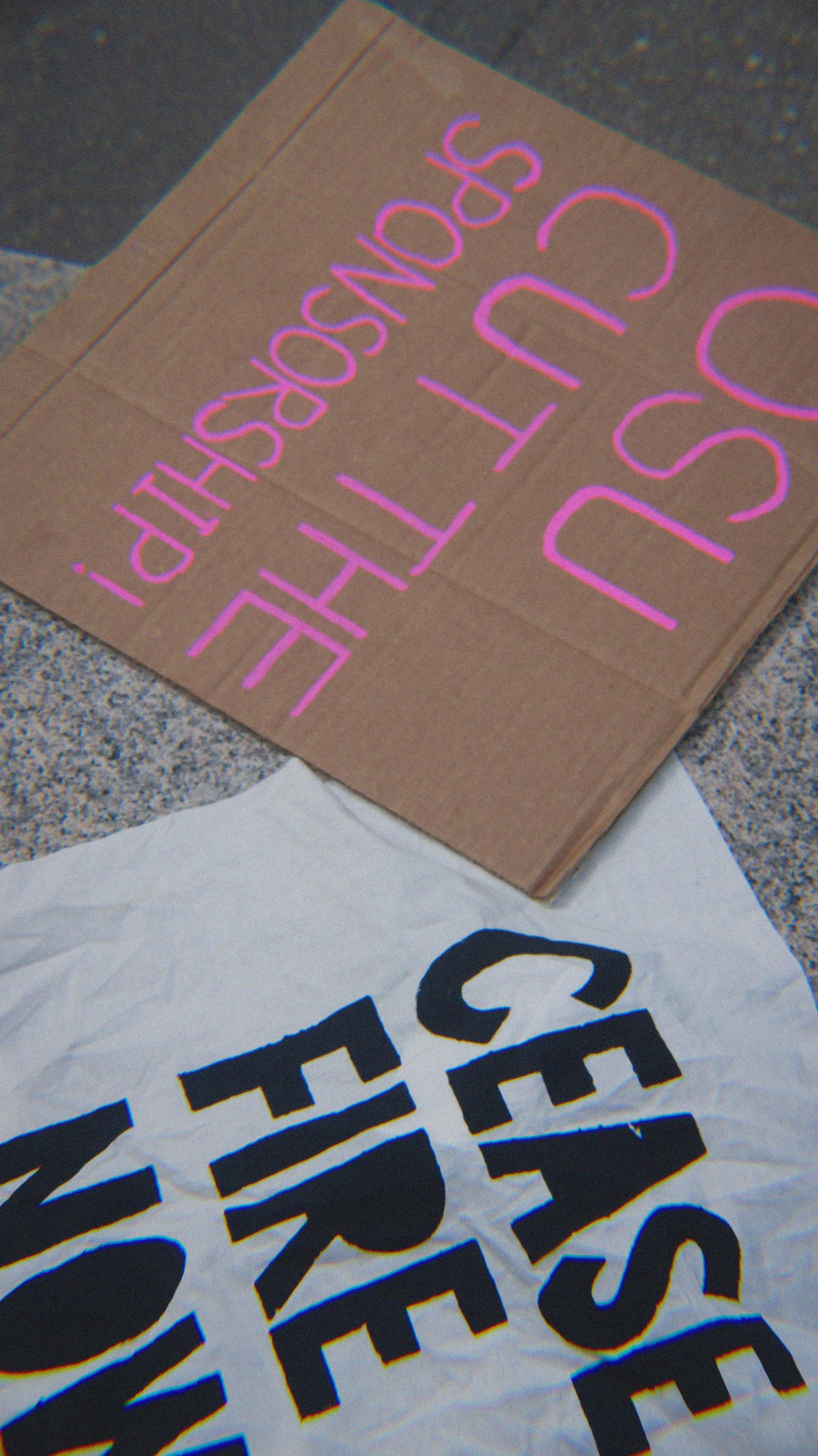 A cardboard sign with purple glowing text that reads: 'THE LOSSorship?'. Partially visible below is a white paper with black text and a logo.