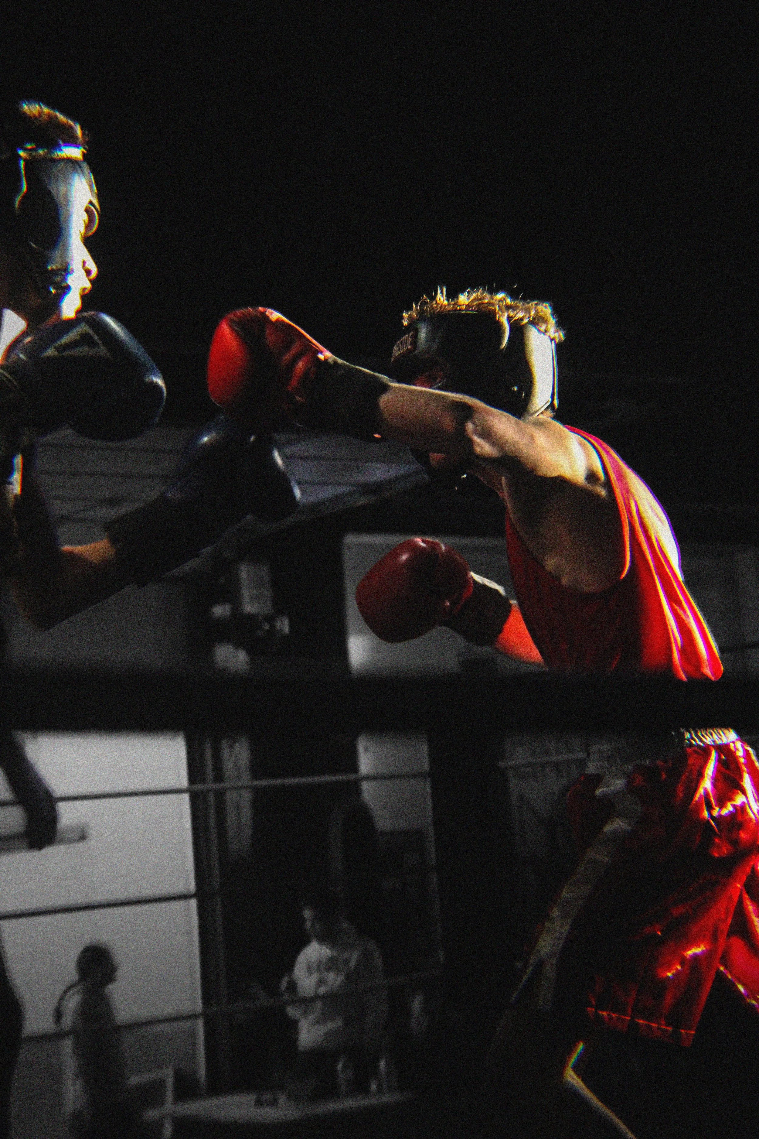 Two women engaged in a boxing match, one in red gloves and the other in black gloves, inside a boxing ring with a black background and spectators watching.