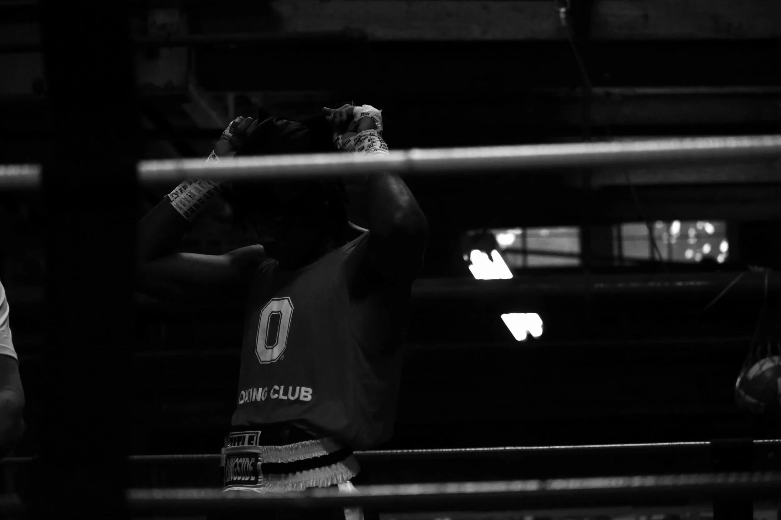 A person wearing boxing gloves and sports gear, adjusting gloves inside a boxing ring, with partial view of a boxing club shirt visible.