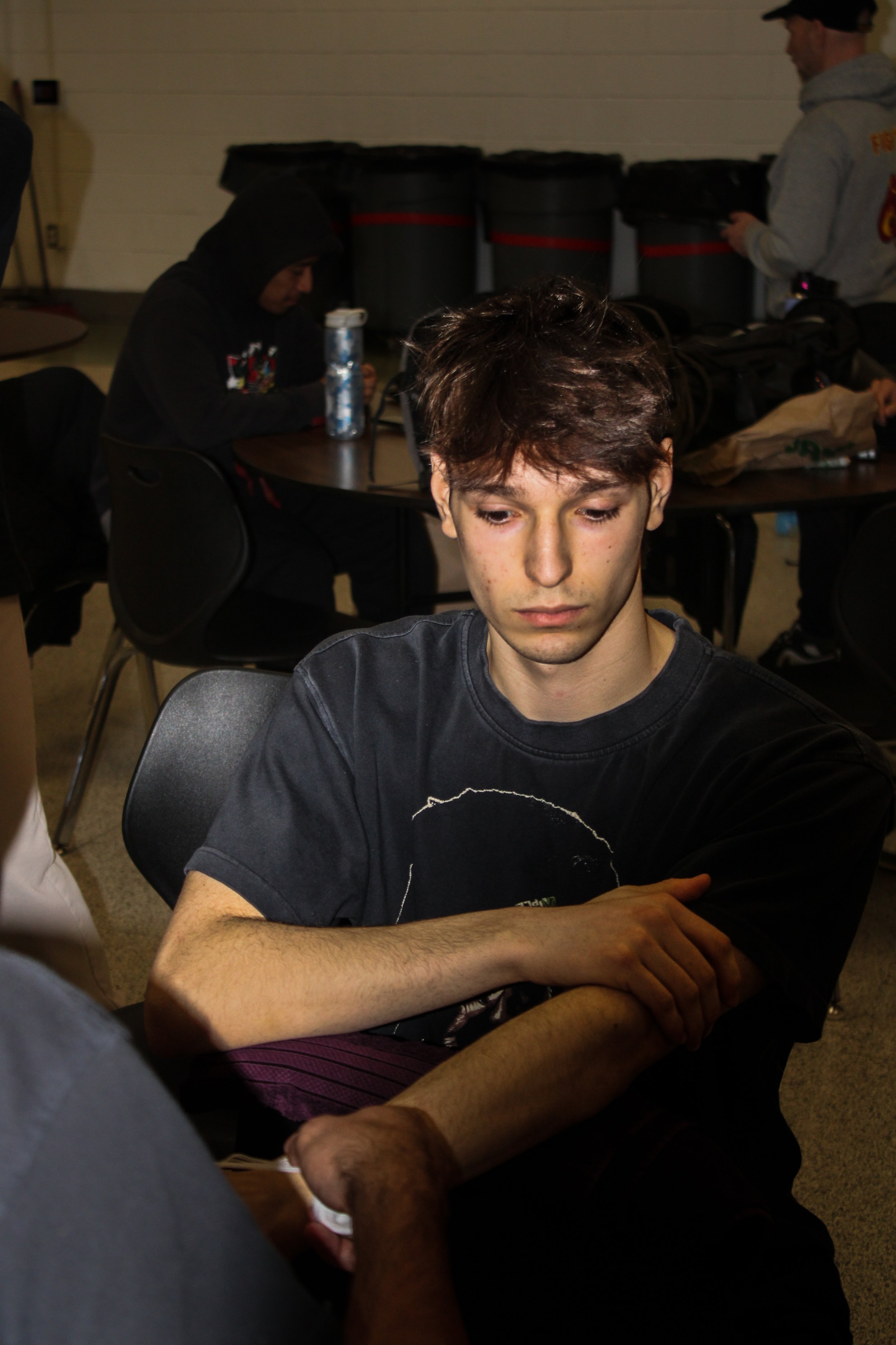 A young man with brown hair and a black T-shirt sitting at a table, with a person in a hoodie and a man in a gray hoodie standing behind him in a room with trash cans in the background.