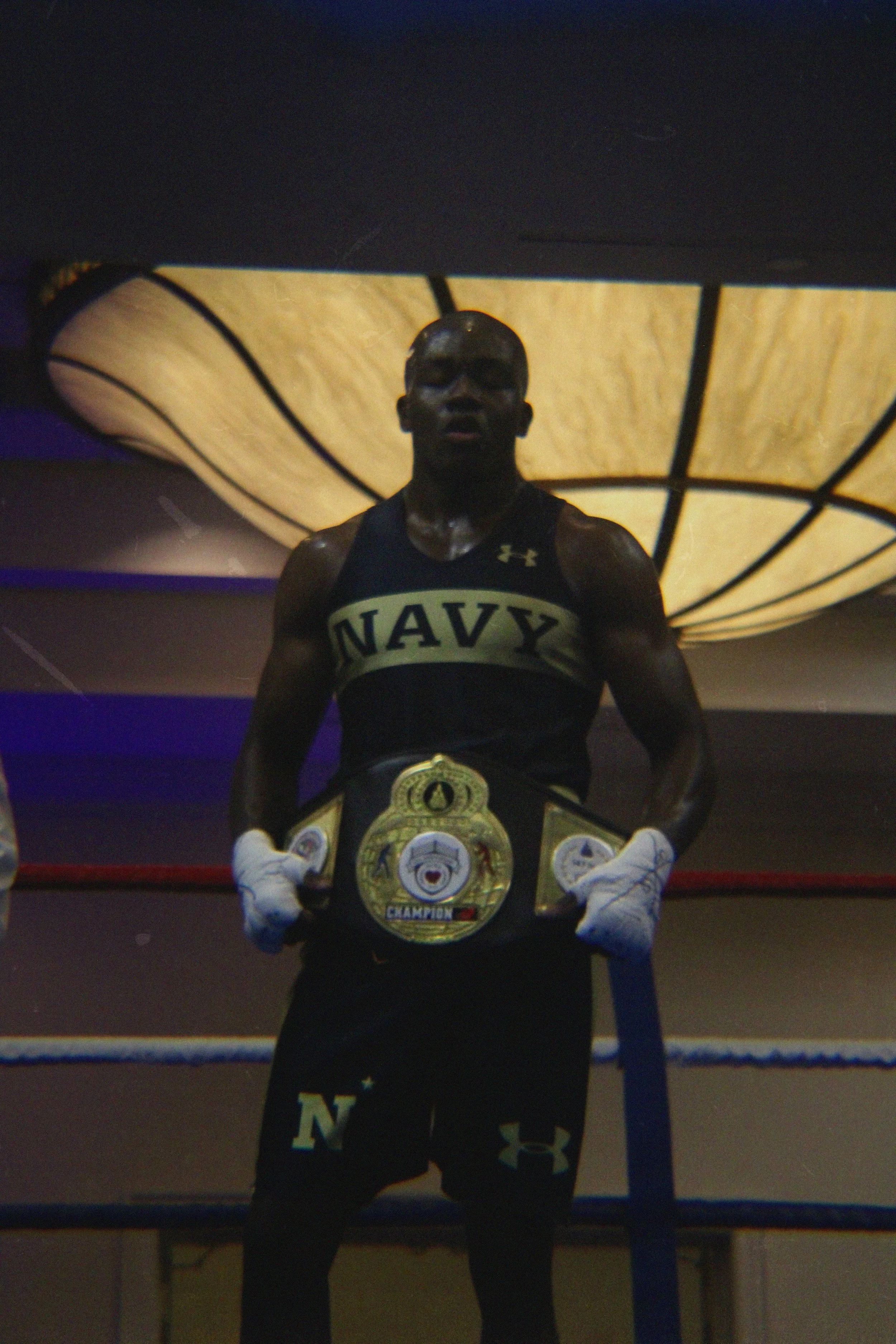 A male boxer with a muscular build, wearing boxing gloves and shorts, standing inside a boxing ring with championship belts hanging around his neck. The background features a large, illuminated ceiling structure and a boxing gym environment.
