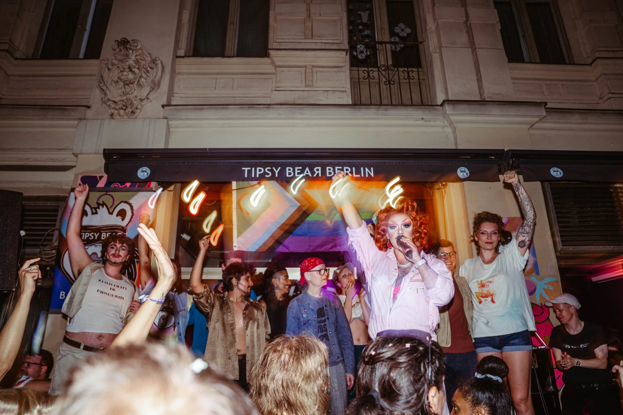 People celebrating at an event with rainbow-colored decorations, a person with bright orange hair speaking into a microphone, and a sign displaying 'Tipsy Bear Berlin' above the crowd.