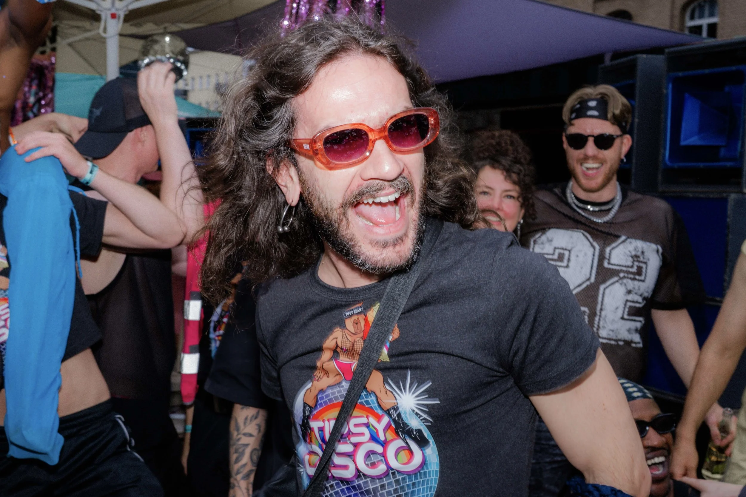 Man with long curly hair and sunglasses, laughing, wearing a black T-shirt with rainbow and unicorn graphic, surrounded by smiling people at an outdoor event.