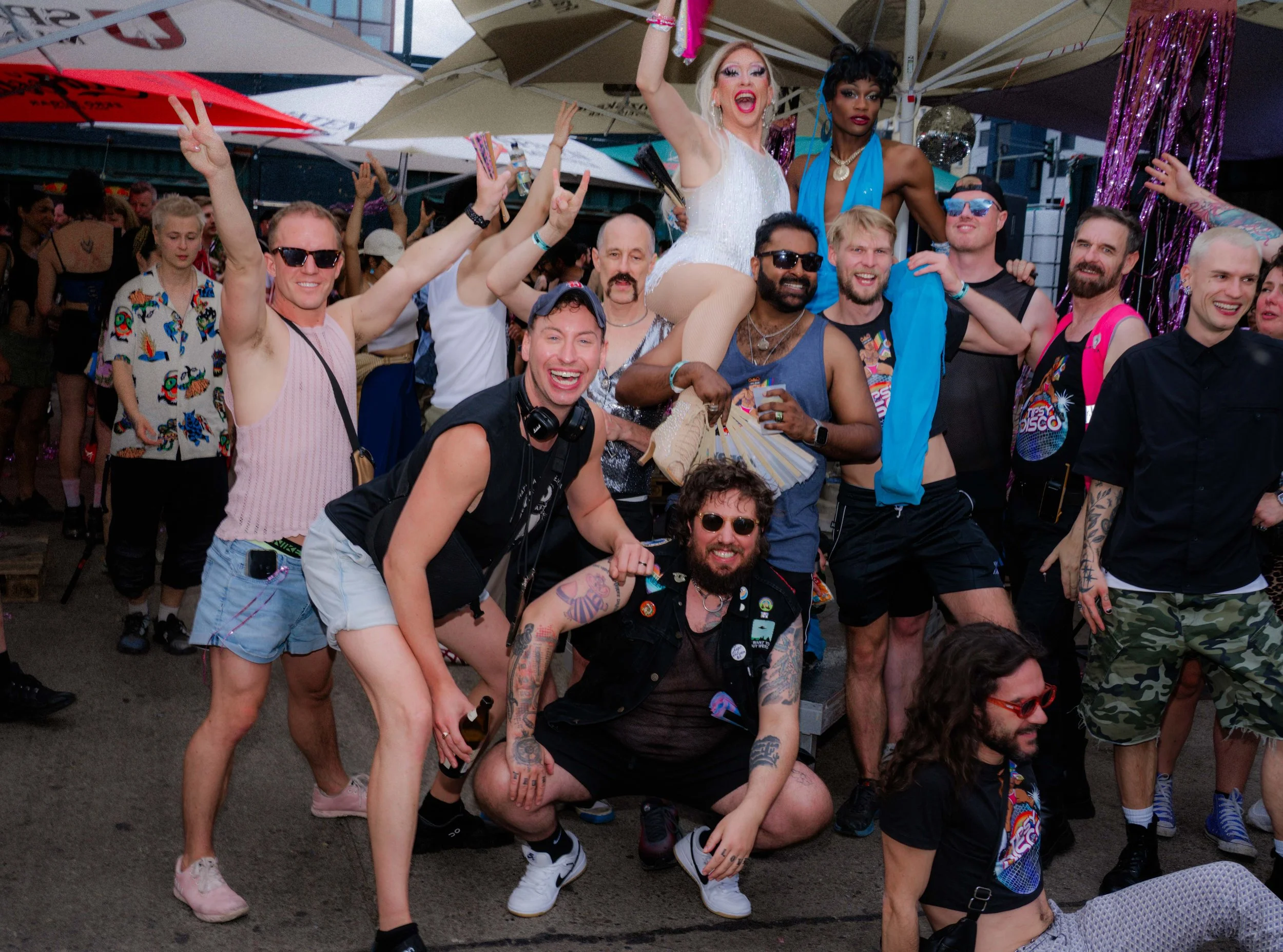 A large group of diverse people at a pride event, with two drag queens on shoulders, celebrating under umbrellas on a city street.