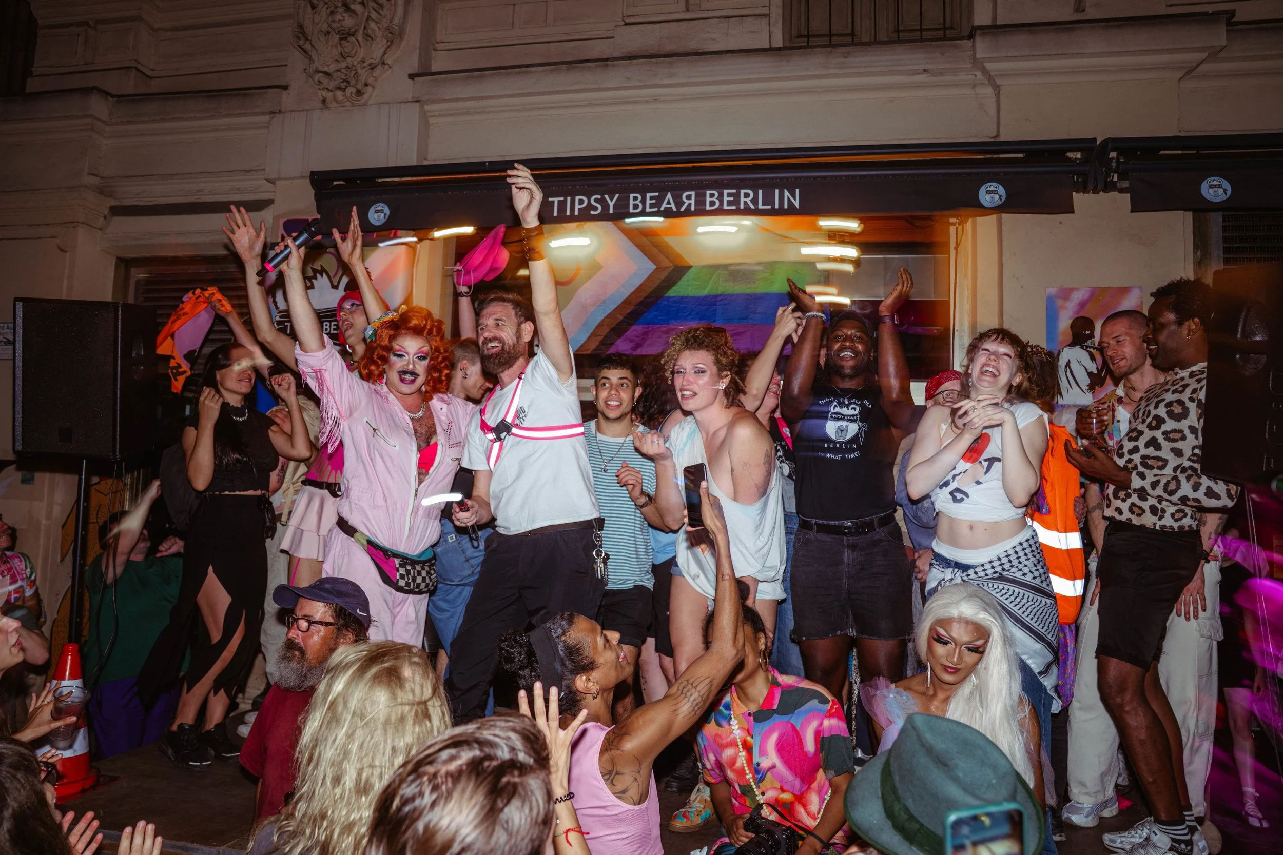 People dancing and celebrating at an indoor event with rainbow colors on a banner in the background