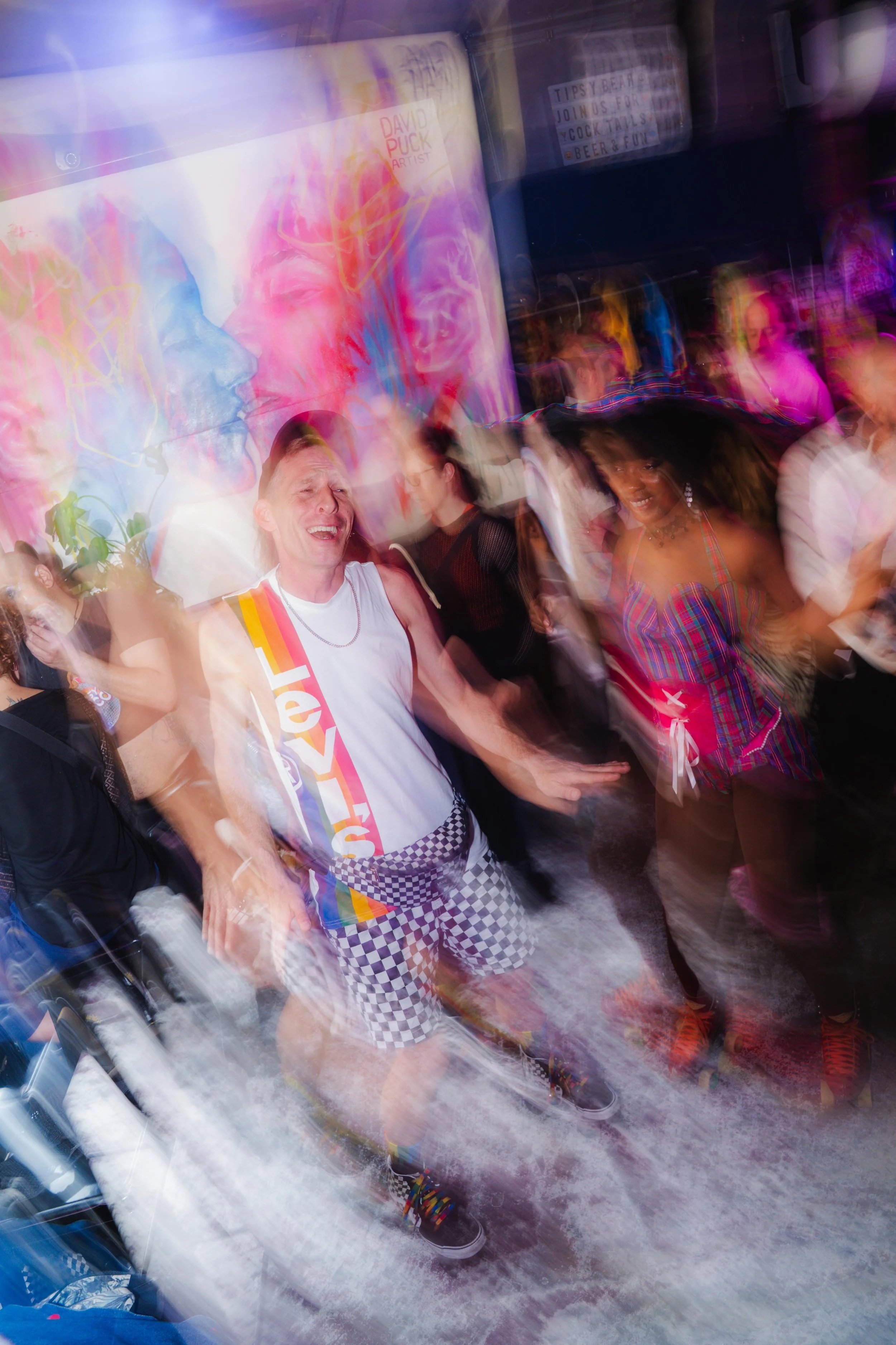 People dancing and smiling at a lively party or event, with colorful artwork and neon signs in the background, motion blur creating a dynamic atmosphere.