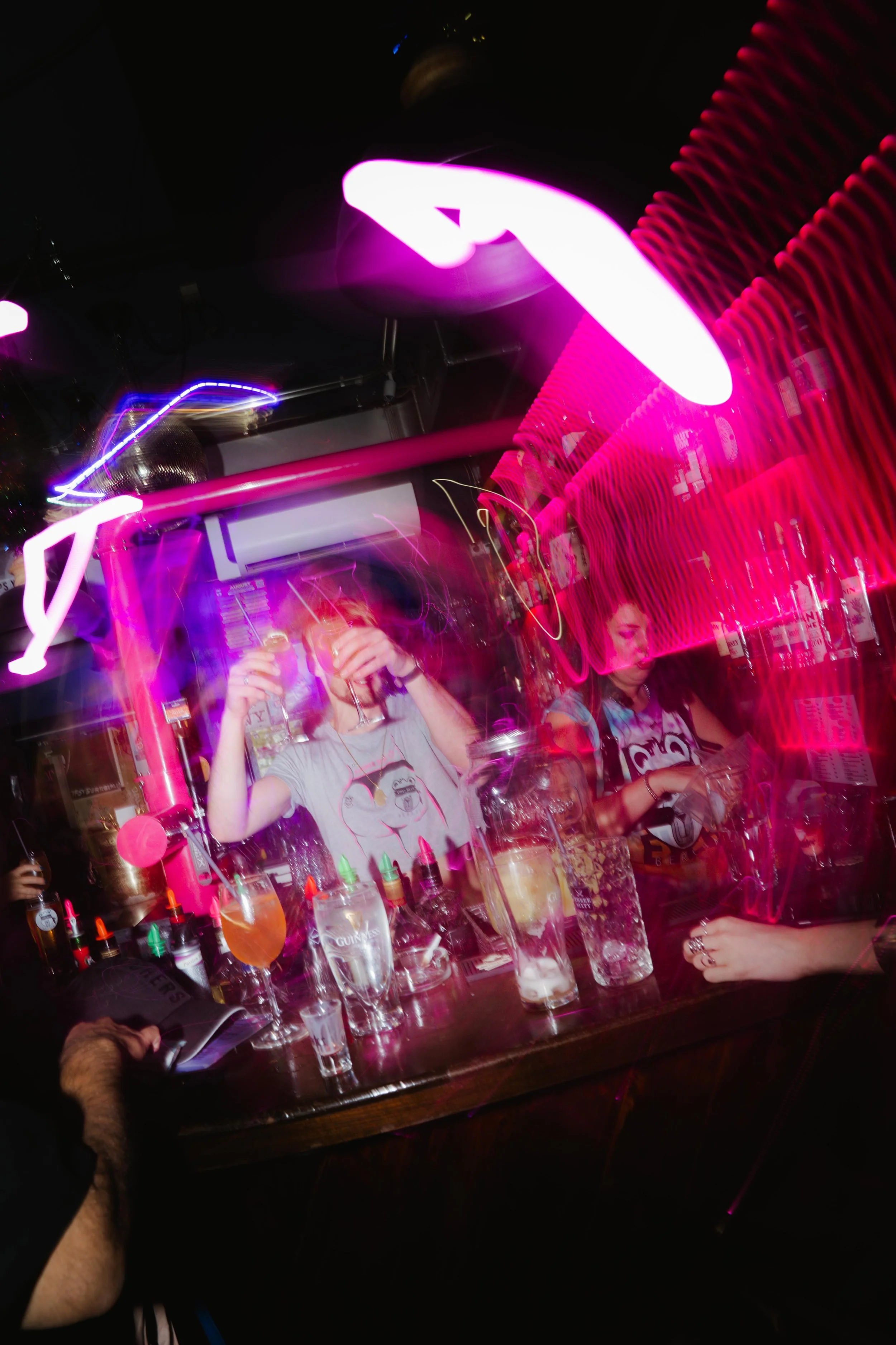 A lively bar scene with neon pink and purple lights, two women standing behind the bar preparing drinks, various bottles and cocktail ingredients on the counter, and drinks in glasses in the foreground.