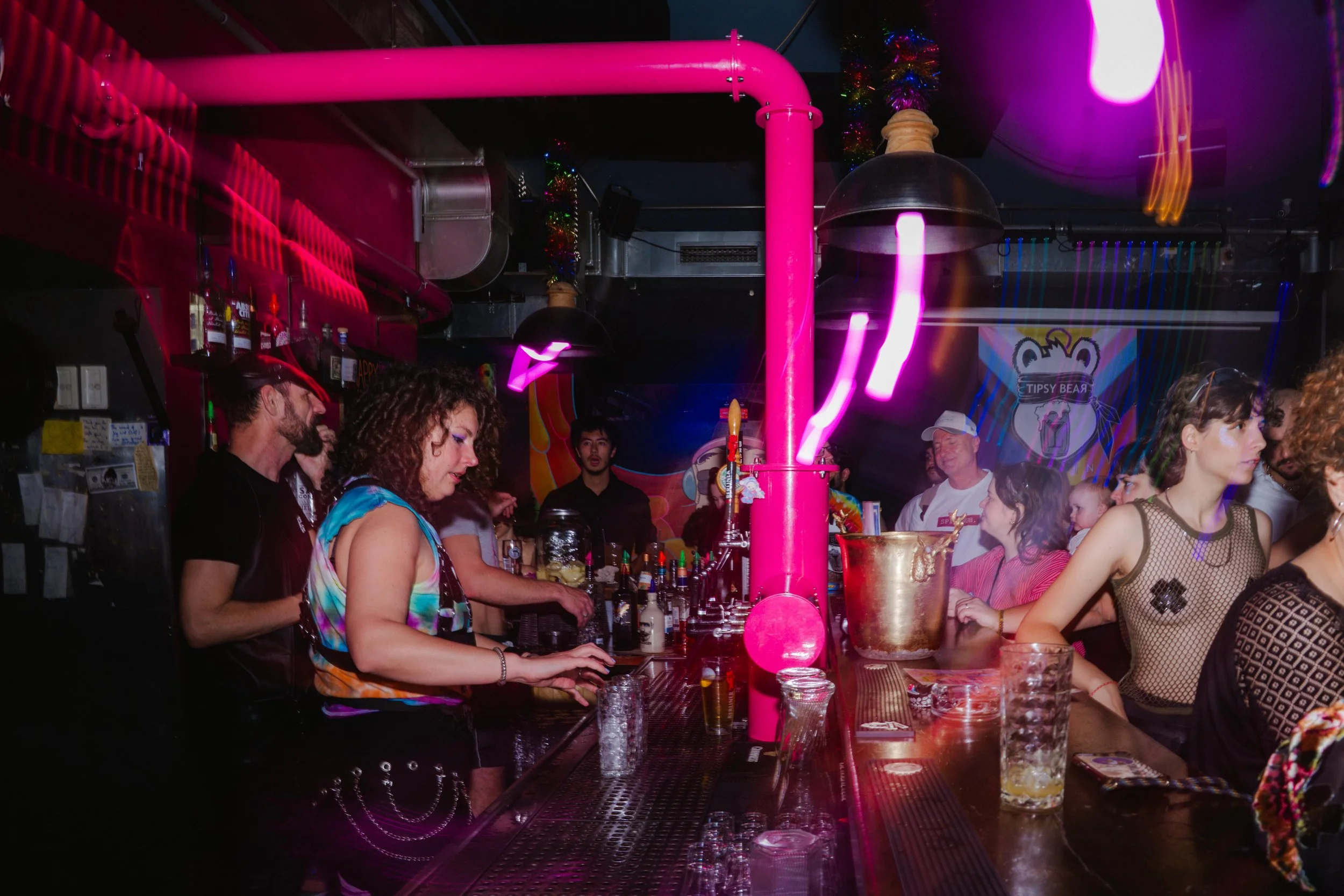 People standing at a bar counter inside a nightclub with colorful lighting, bottles, glasses, and a pink pipe in the foreground.