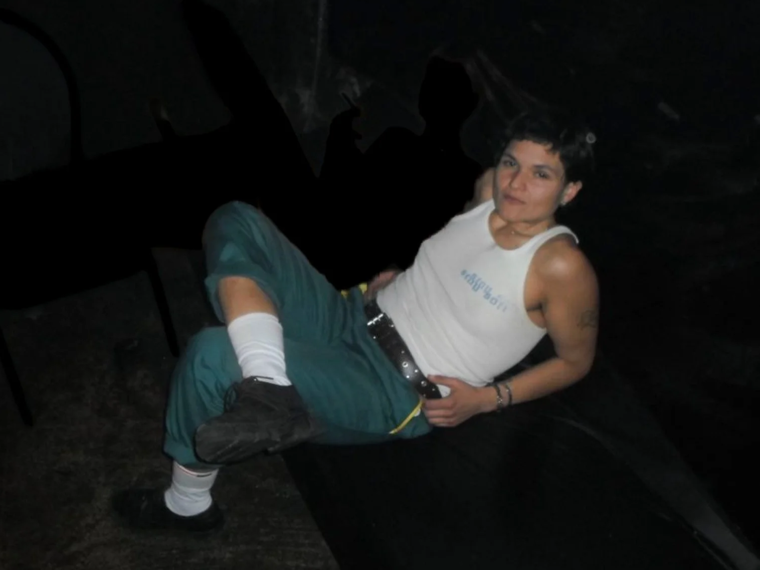 A person with short dark hair and tattoos on both arms is lying on a black surface in a dark room. They are wearing a white sleeveless shirt, teal shorts, a black belt, white socks, and black shoes. They are positioned with one arm resting on their stomach and the other behind their head, giving a slight smile.