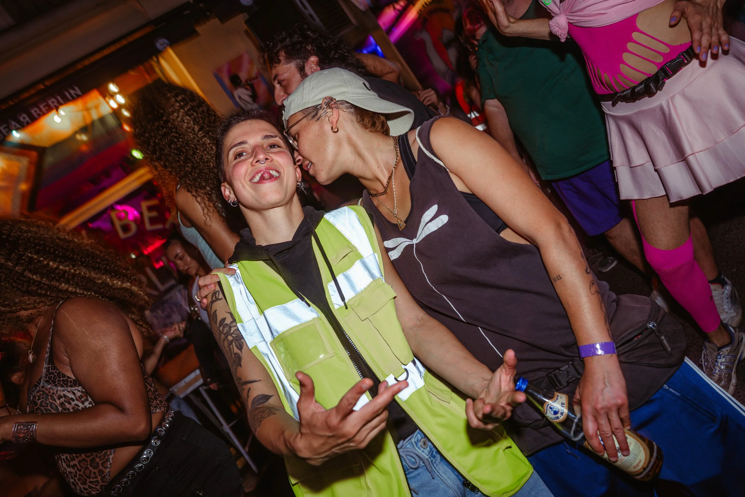 People socializing at a lively indoor event, with a woman in a high-visibility vest and others dressed in casual or eccentric clothing, some with tattoos and piercings, under colorful lighting.