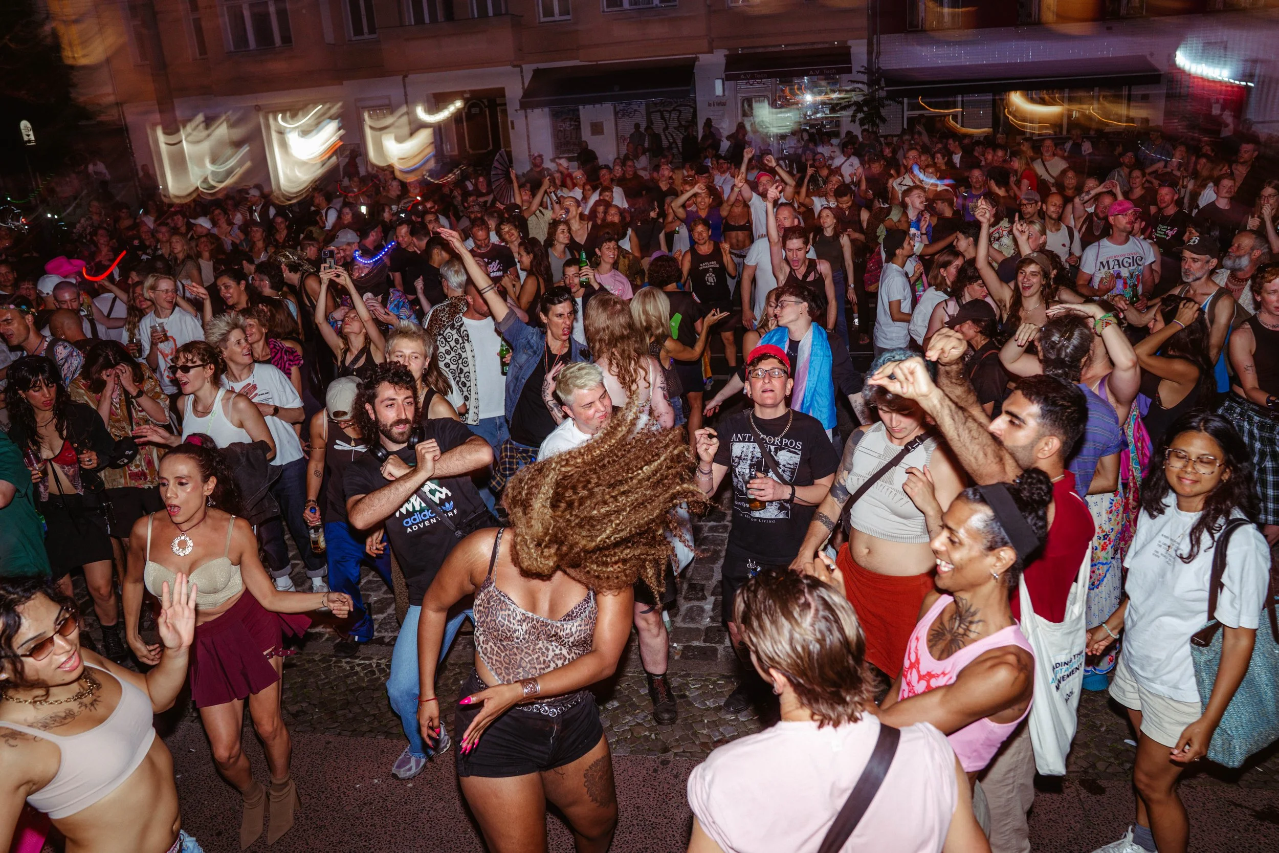 Crowd of people dancing and enjoying music outdoors at night, some with drinks, in a lively street scene.
