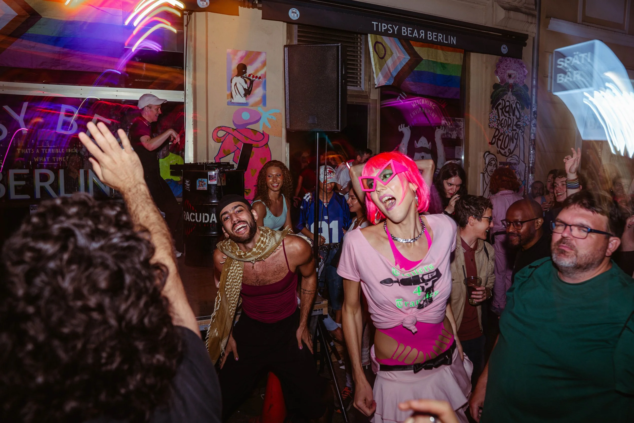 Crowd of people dancing and smiling at a lively party or dance event in a colorful, vibrant setting with graffiti art and bright lighting.