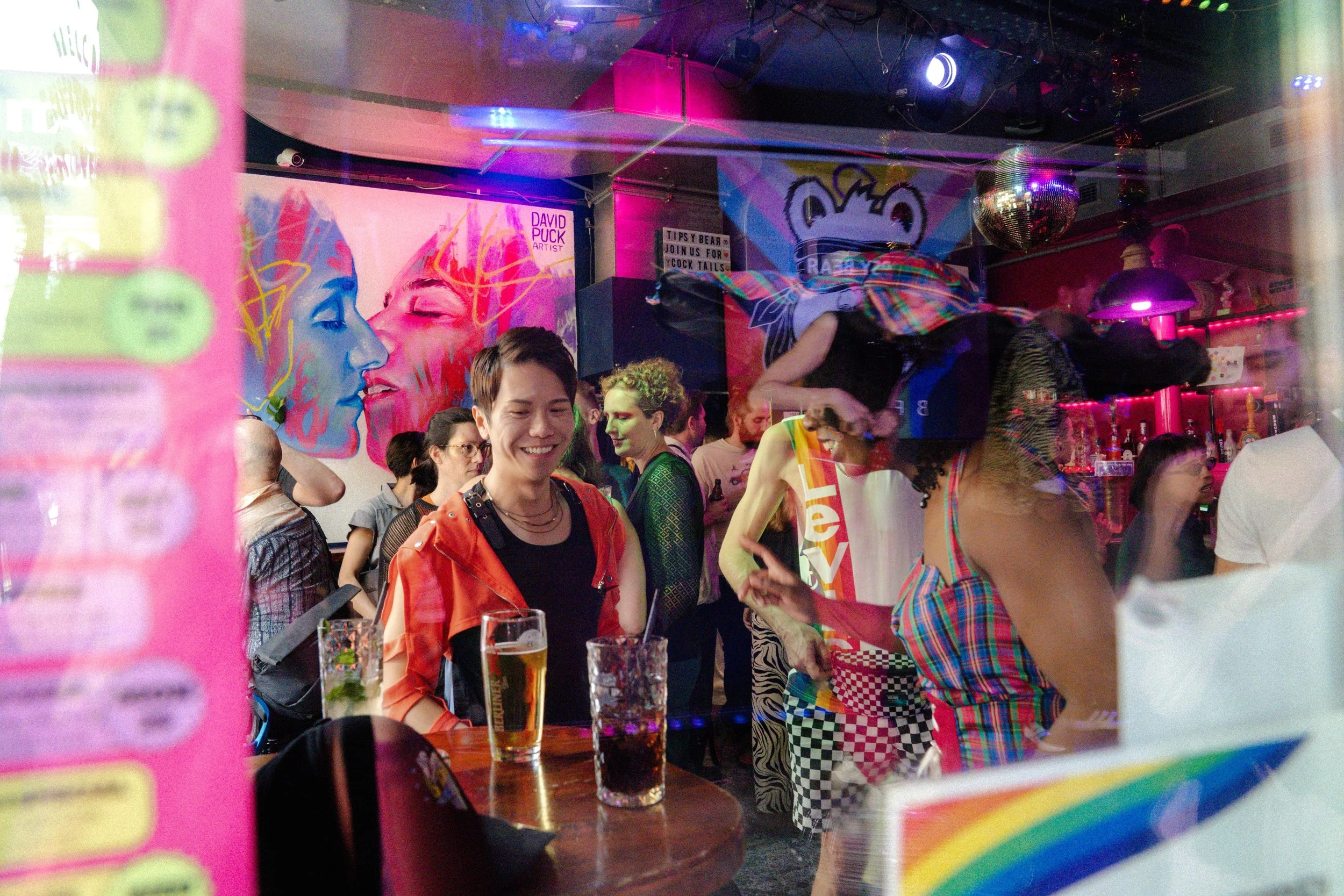 People dancing and socializing in a colorful nightclub or bar with vibrant lighting, a large mural of two faces, and a disco ball hanging from the ceiling.