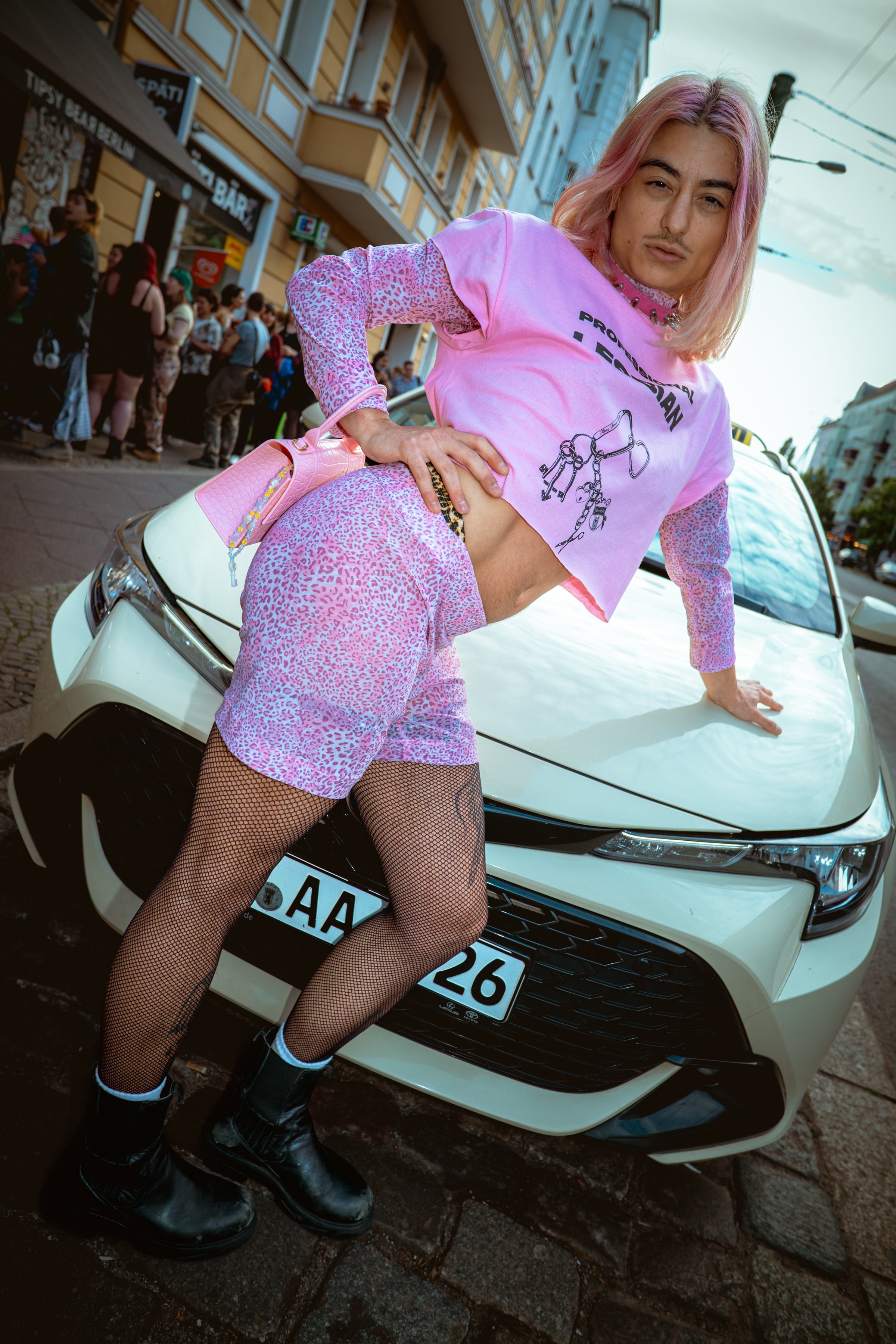 A woman with pink hair is posing on the hood of a white car on a city street. She is wearing a pink graphic shirt, matching pink leopard print shorts, fishnet stockings, and black boots. In the background, there is a crowd of people in line outside a building with a yellow facade.