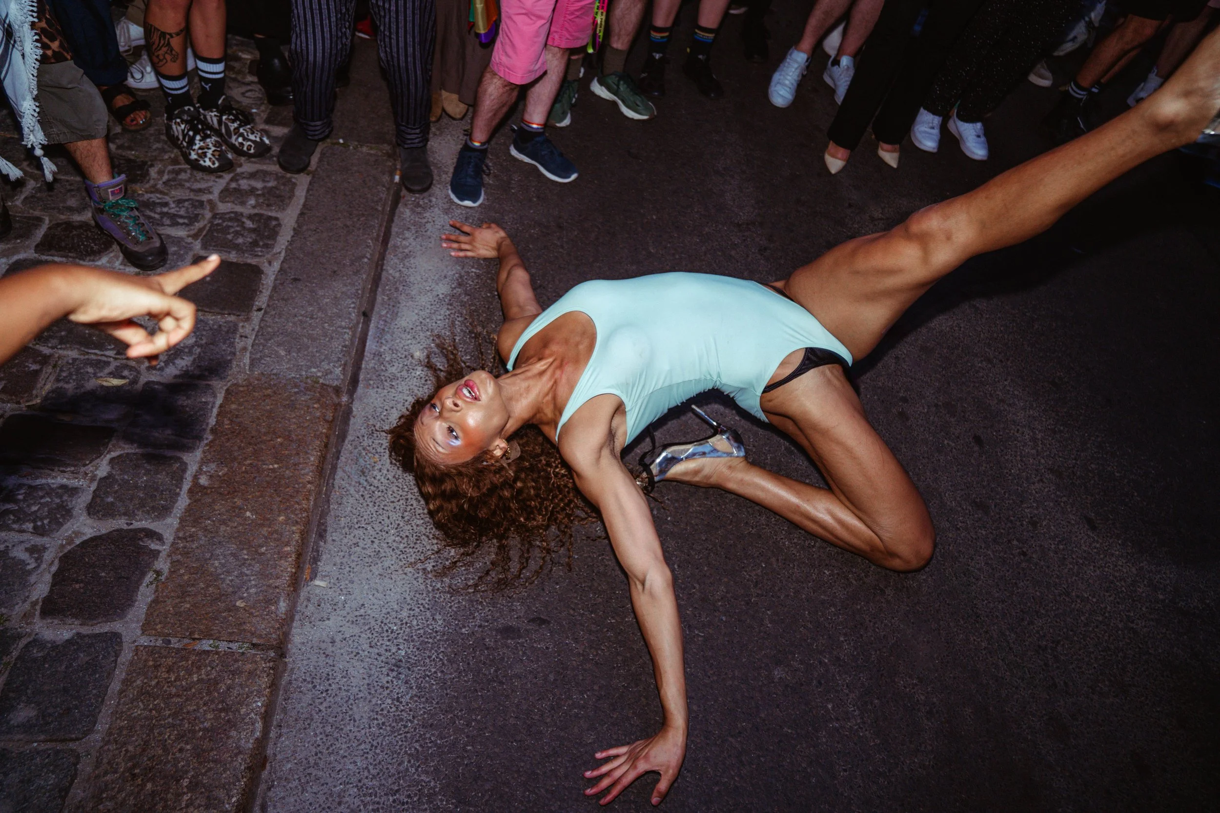 Woman laying on the ground with her side on the cobblestone street, surrounded by a crowd of people standing around her.