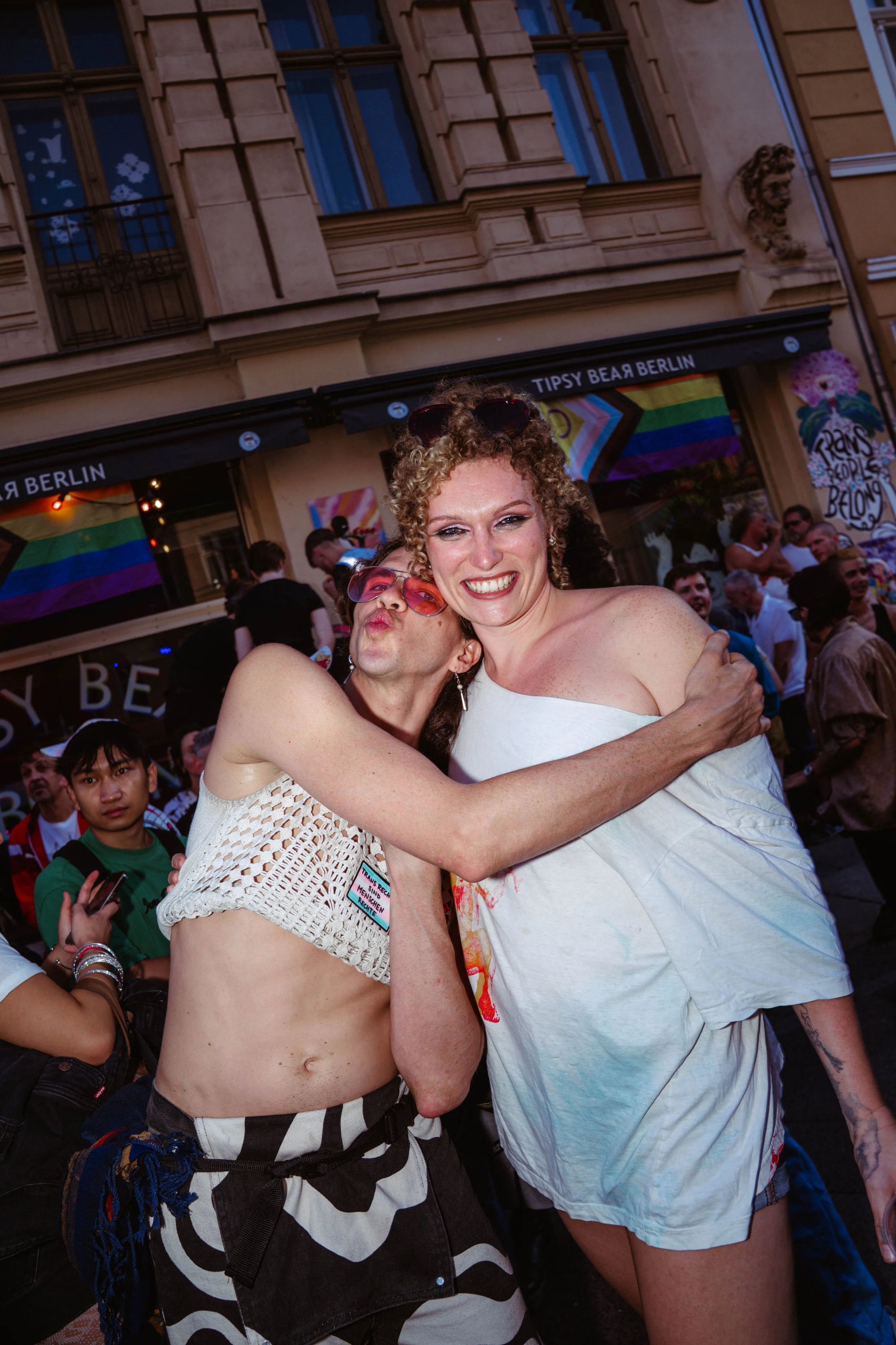 Two smiling women hugging each other at a pride event, with a crowd and rainbow flags in the background.