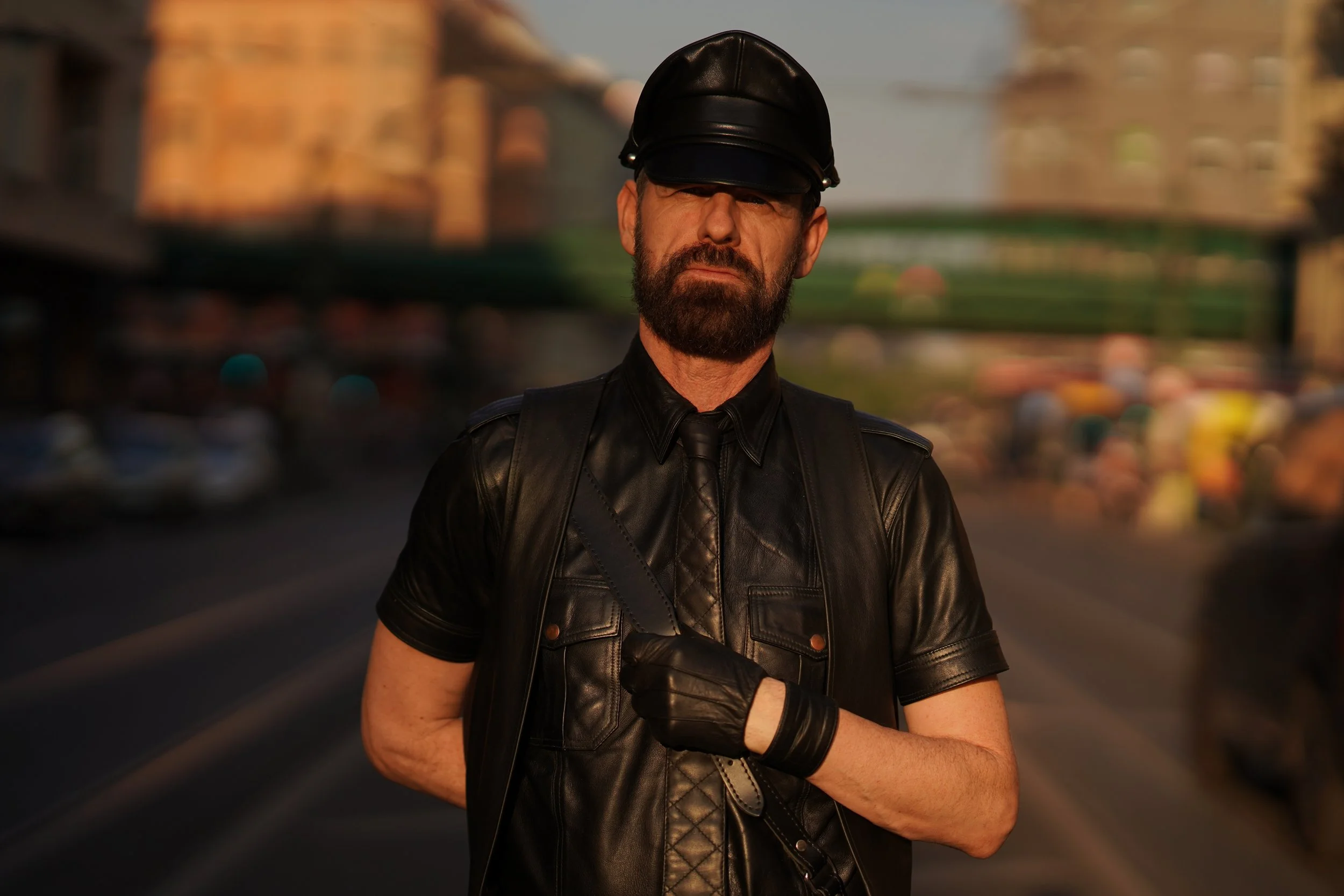 A man with a beard wearing a black leather police officer's hat and leather jacket, standing on a city street during daylight hours.