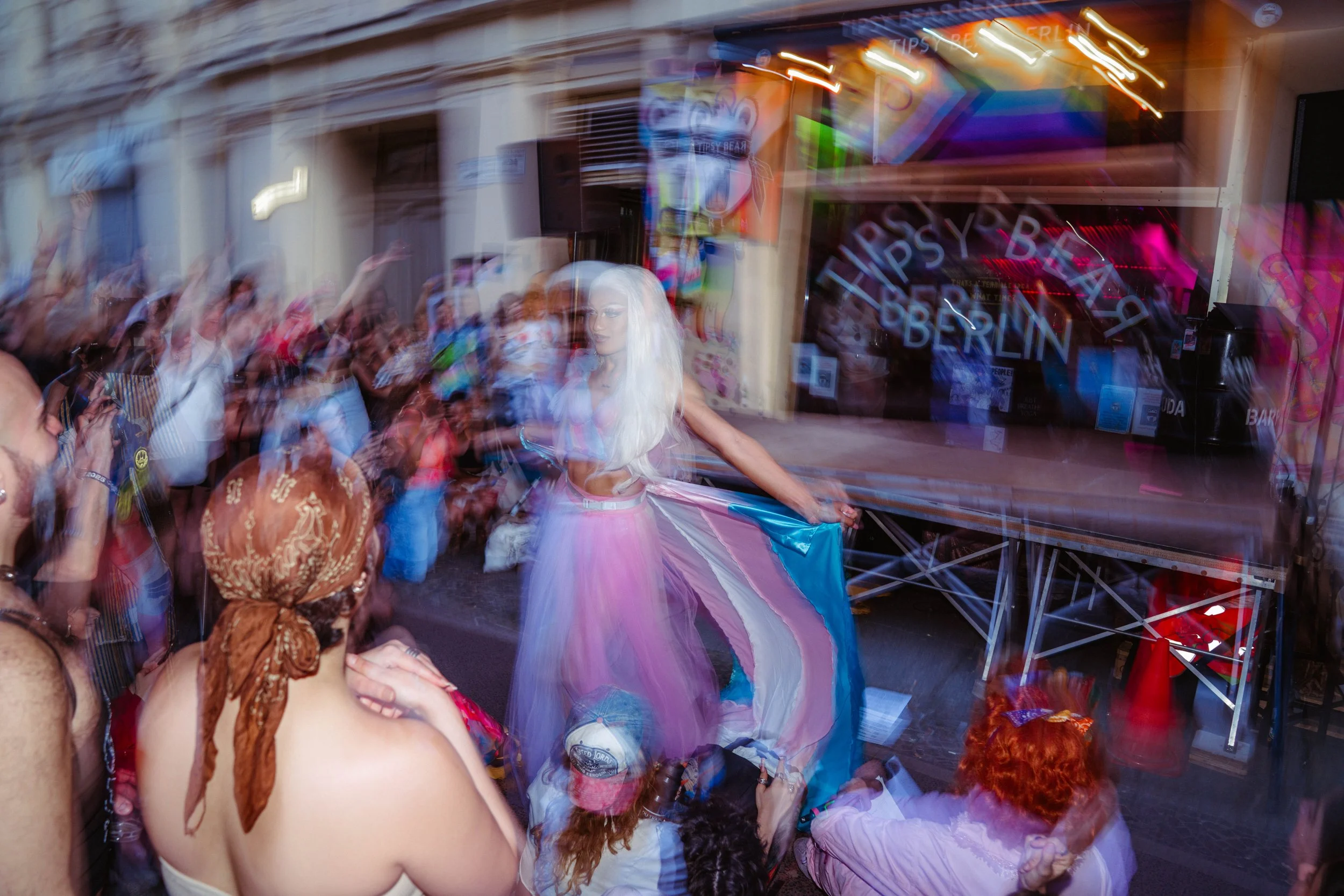 A crowd of people gathered outdoors at a lively event, with a woman in a pastel-colored dress and blonde hair in the center. Neon signs reflect on glass, including the words 'Tipsy Berlin'.