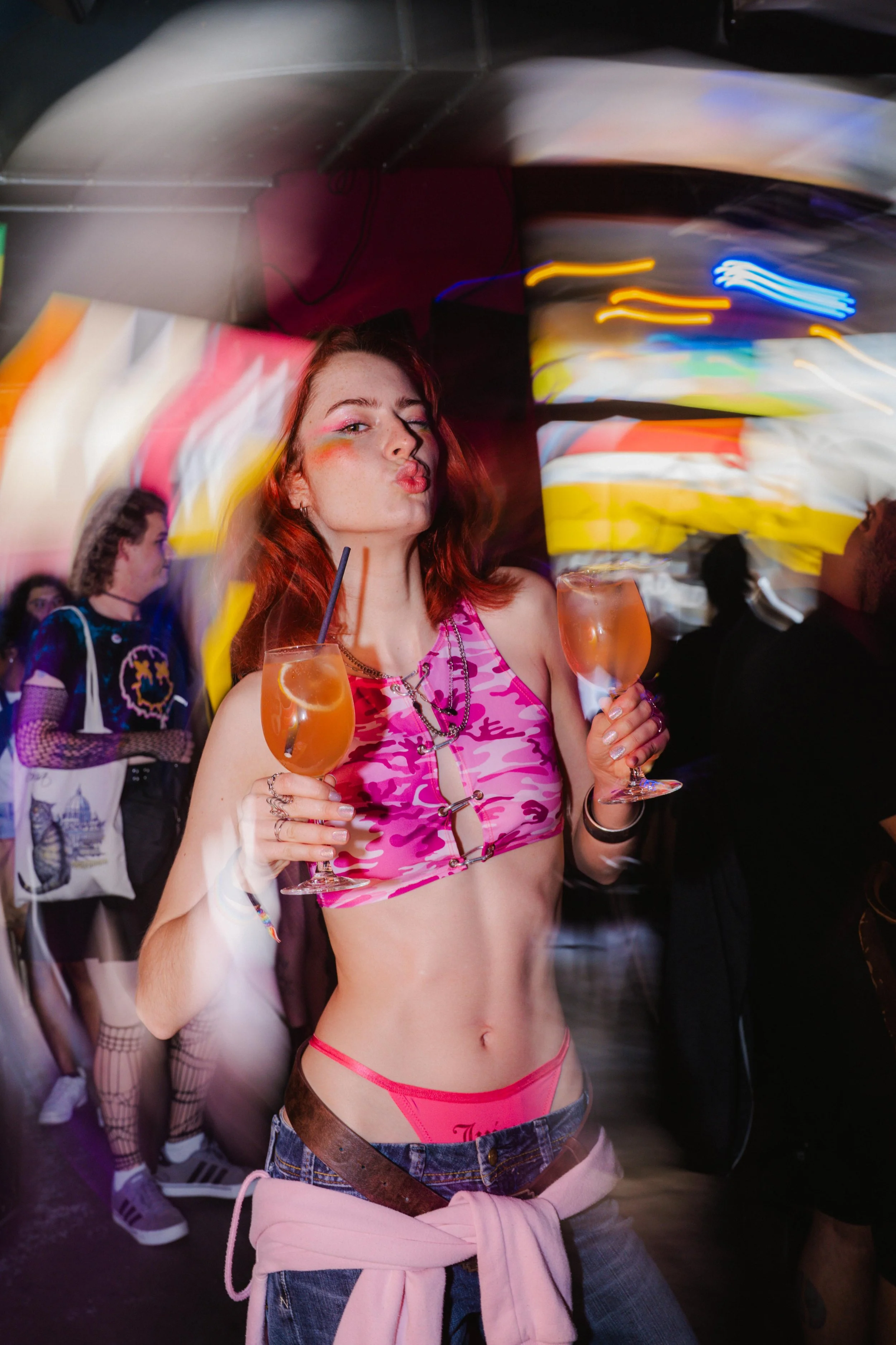 Woman with red hair wearing pink camouflage crop top, pink underwear, and holding two drinks in a club or party setting. Blurred background with other people and colorful lights.